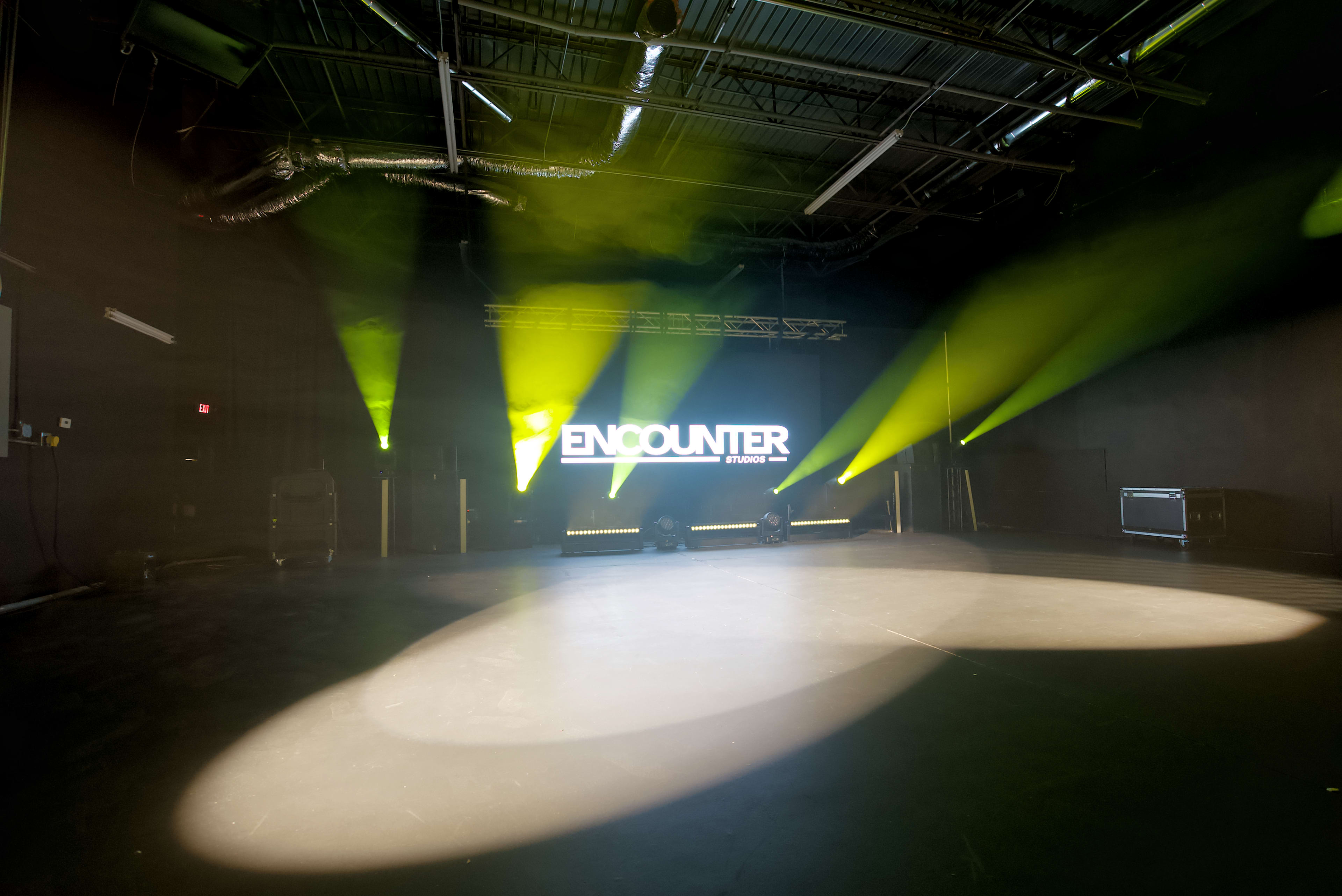The image shows a performance space with a large illuminated sign reading "ENCOUNTER" and focused stage lighting casting beams onto an empty stage.