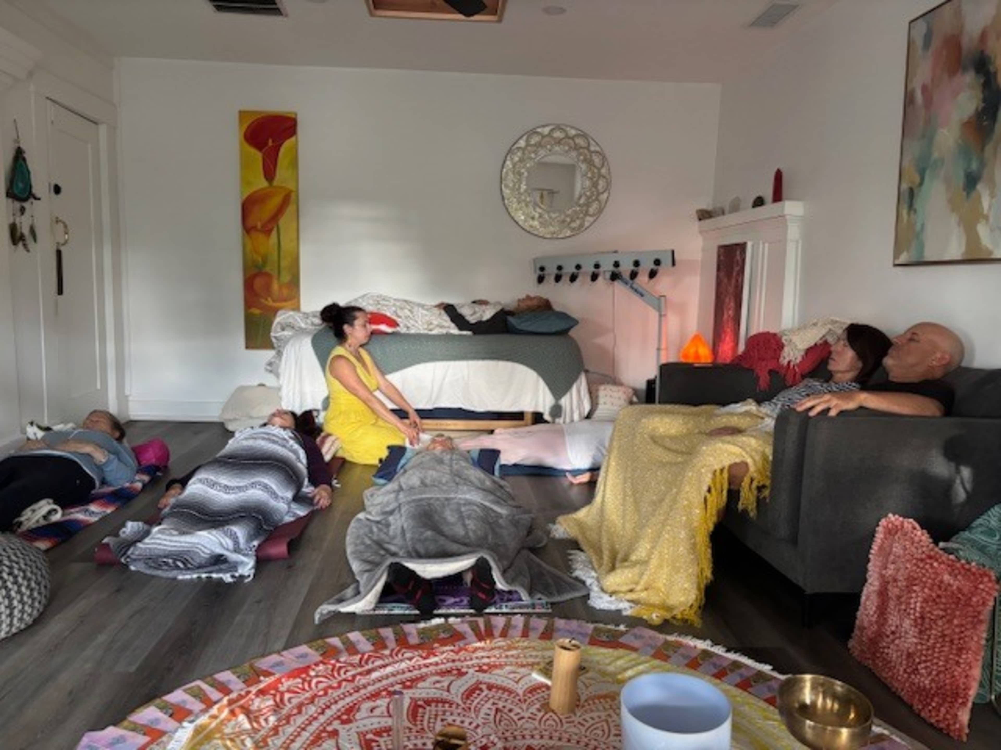 A group of six people is lying on mats in a spacious, brightly lit room with a bed and a couch in the background.