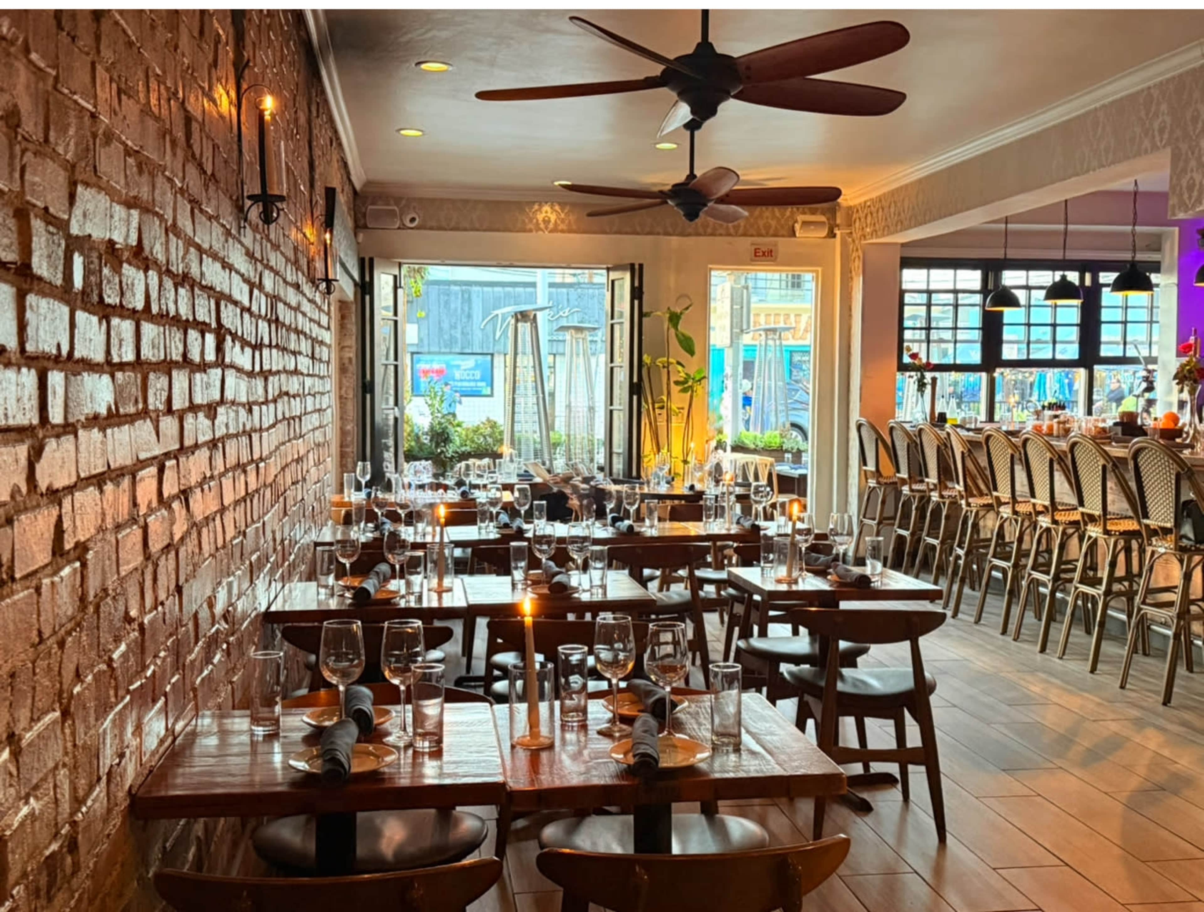 The image shows a restaurant interior featuring rows of wooden tables set with glassware, surrounded by a brick wall and illuminated by warm lighting.