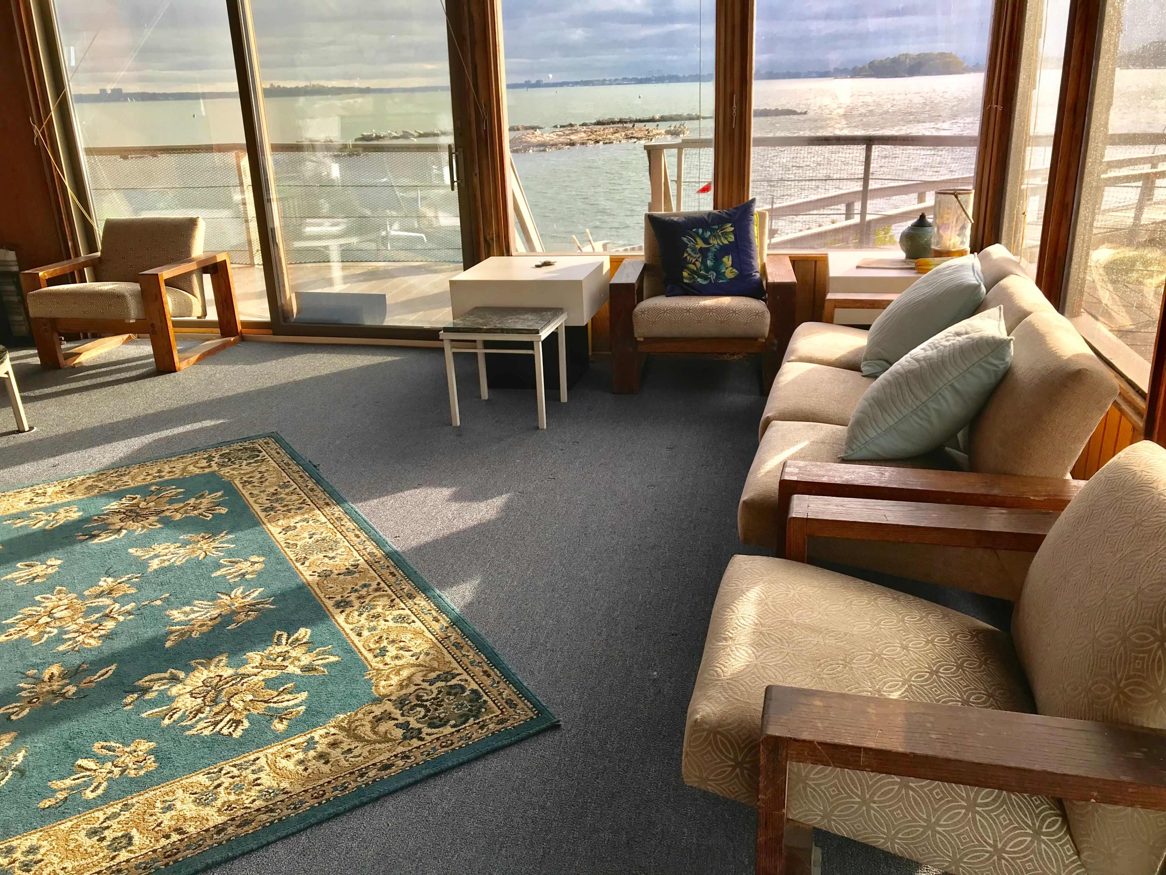 The room features a seating area with a large rug, two armchairs, a sofa, and large windows overlooking a body of water.