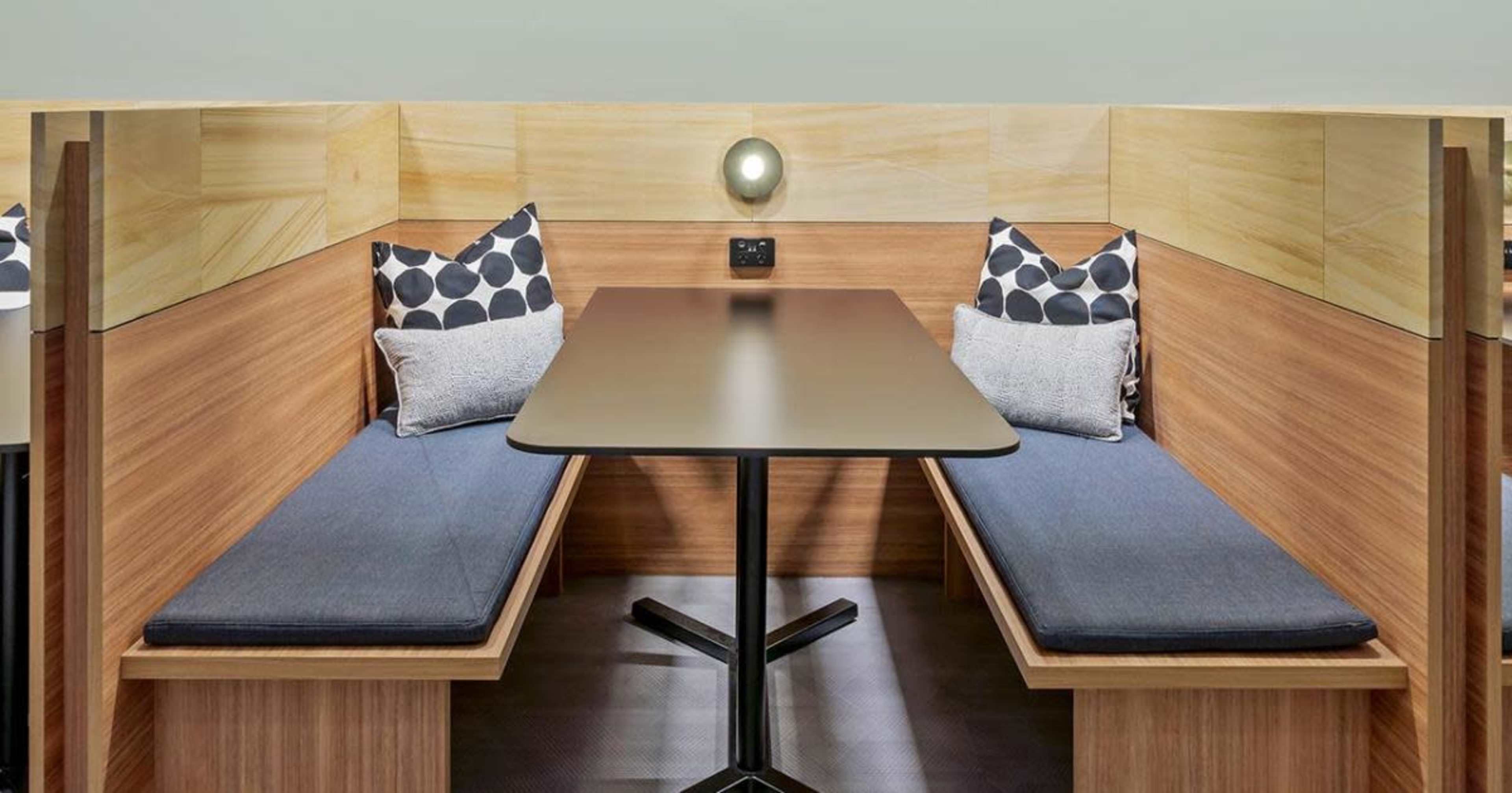 A pair of wooden booth seating areas with a central table, equipped with cushions and a wall-mounted light.