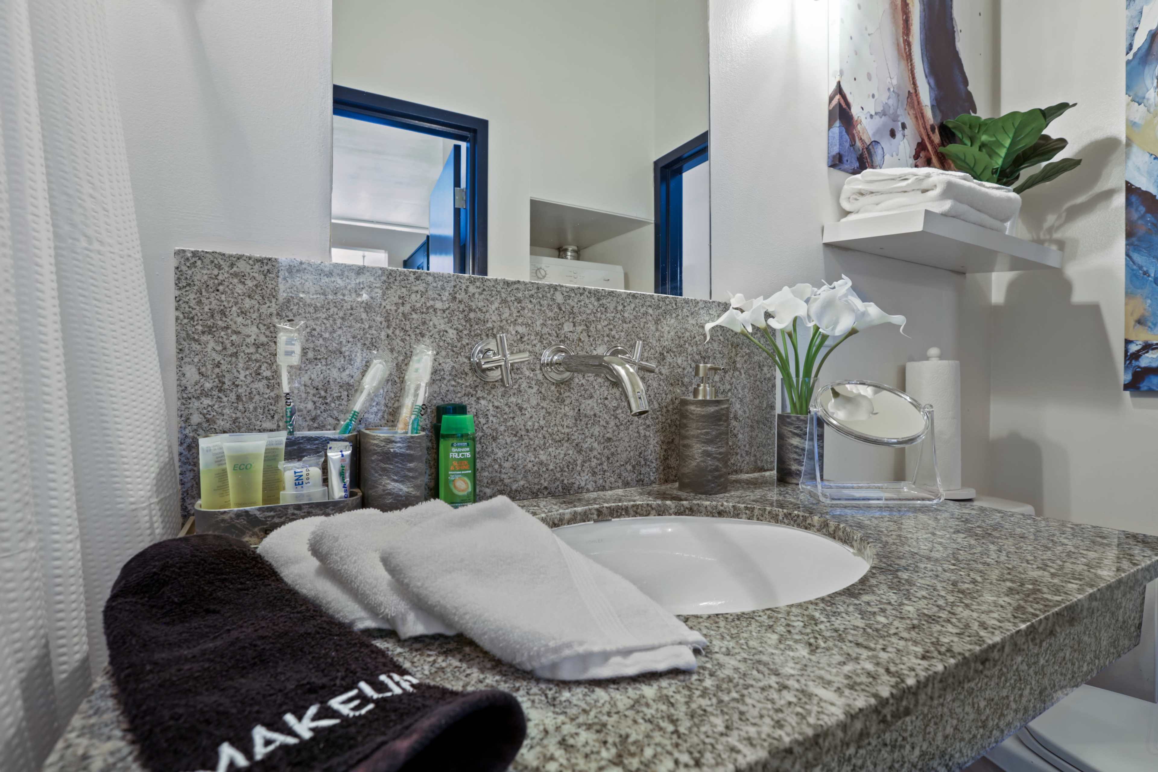 A clean bathroom countertop with a sink, decorative plants, toiletries, a towel, and a mirror reflecting the room.