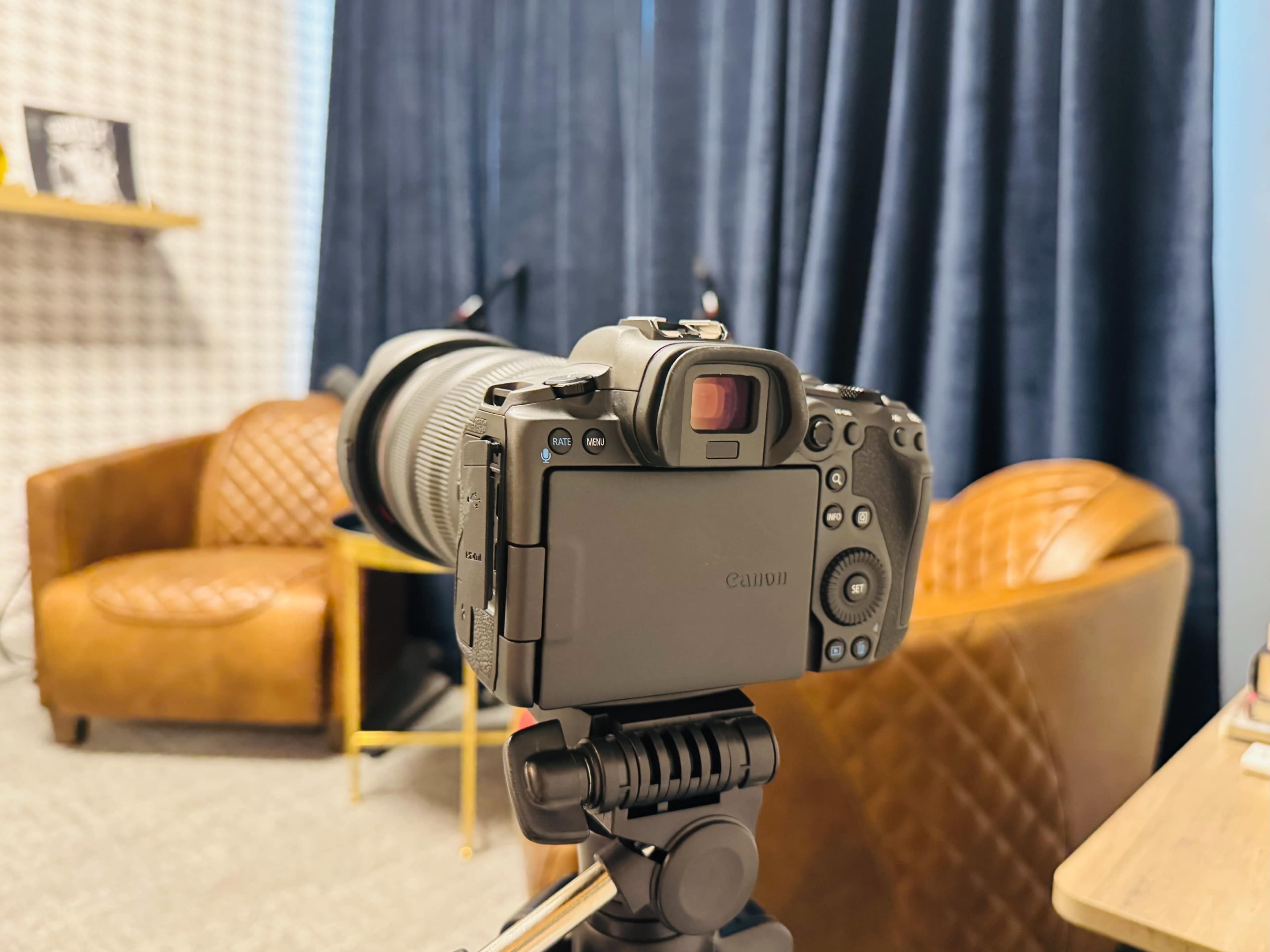 A Canon camera is set up on a tripod, facing two leather armchairs in a room with dark curtains and a patterned wall.