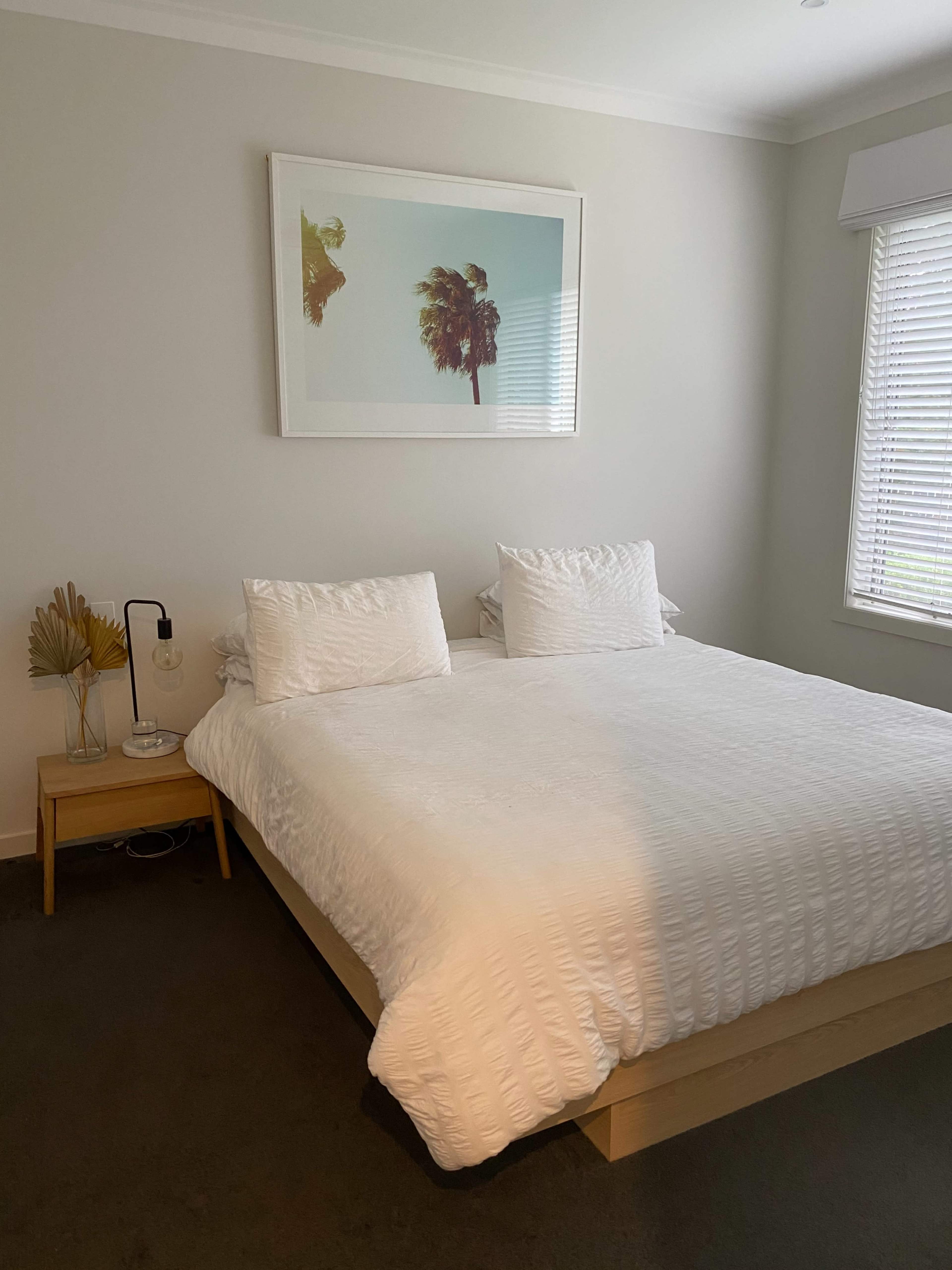 A minimalist bedroom with a large bed, two white pillows, a nightstand, and a framed picture of palm trees on the wall.