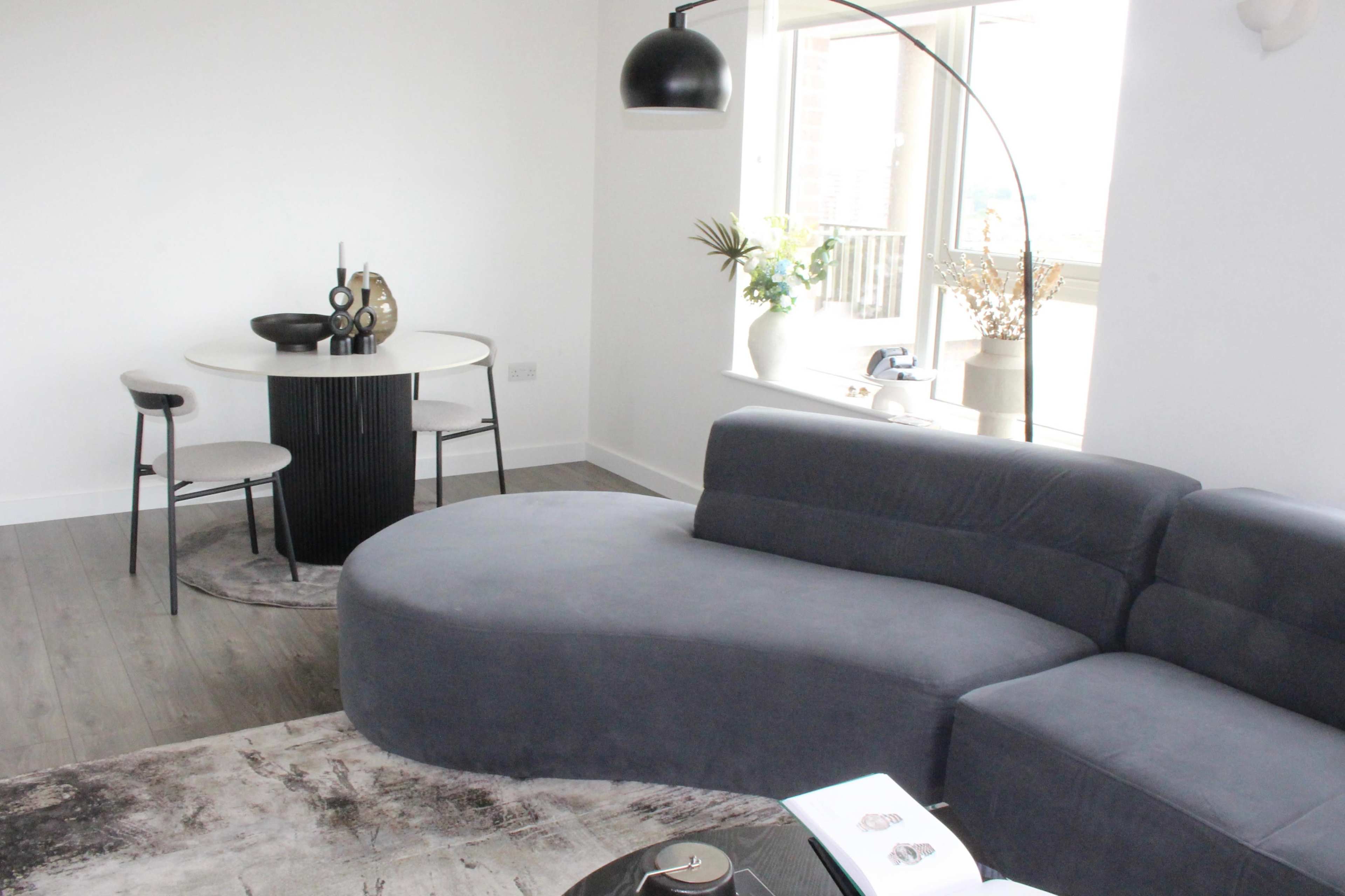 A modern living room features a curved gray sofa, a round dining table with two chairs, and large windows allowing natural light to enter the space.