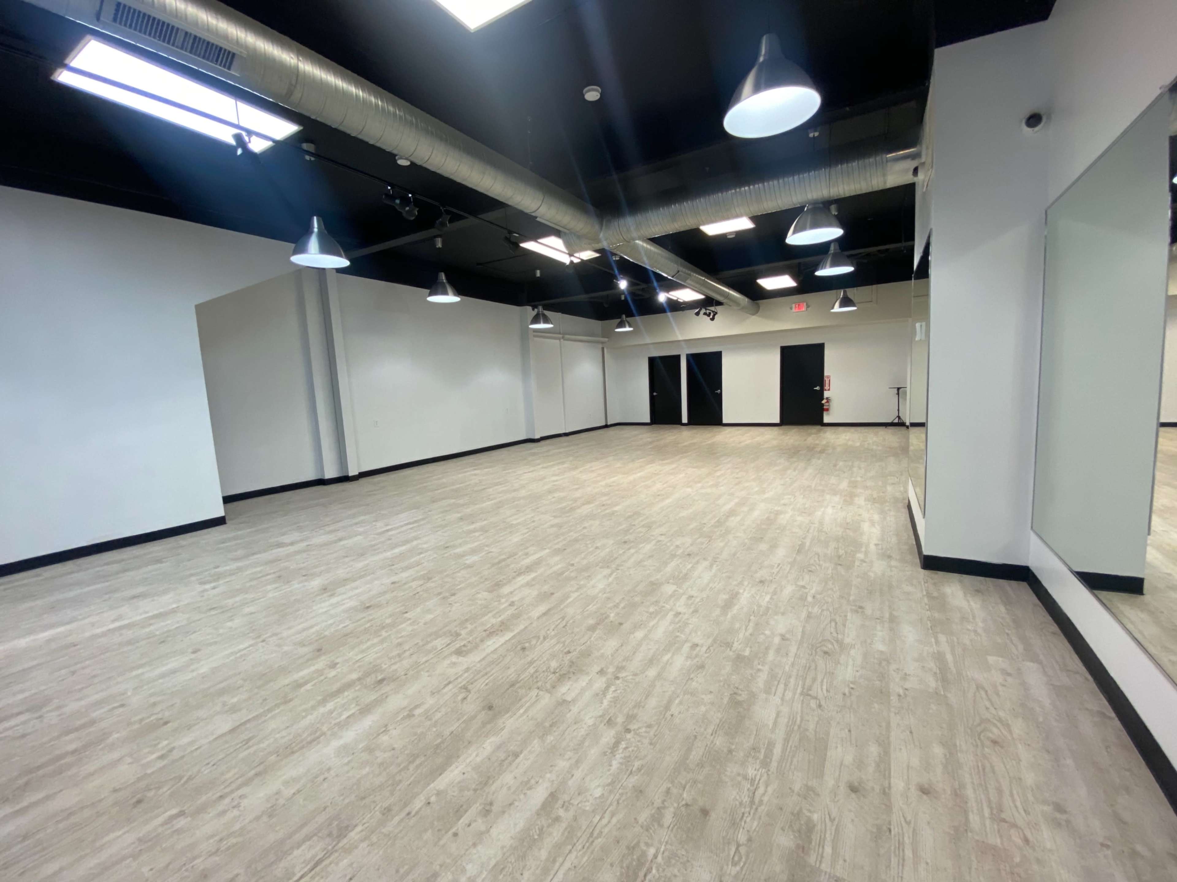 A spacious, empty room with wooden flooring, white walls, and several ceiling lights.