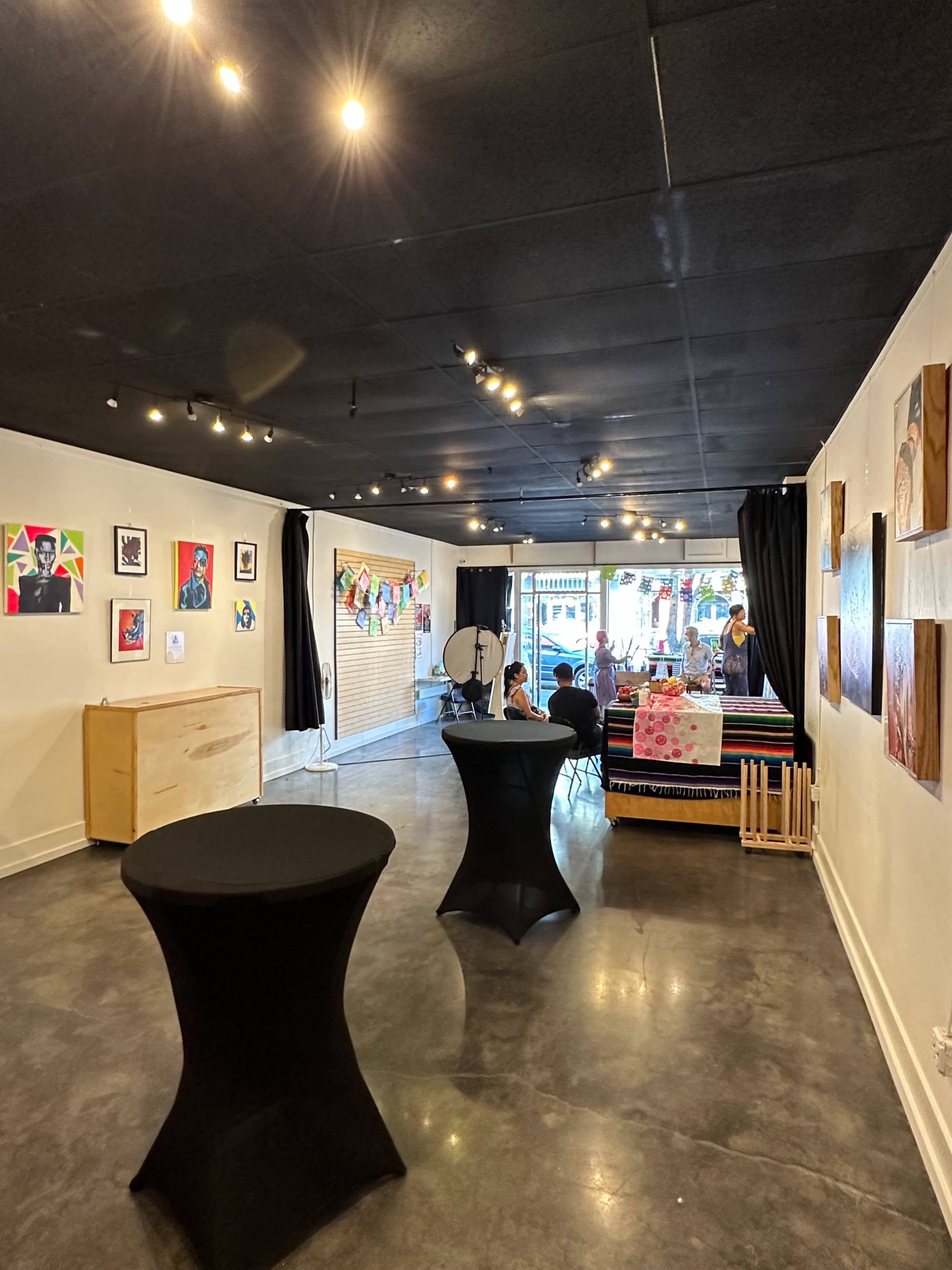 An art gallery with a polished concrete floor, featuring various artworks on the walls and two small, round tables in the foreground.