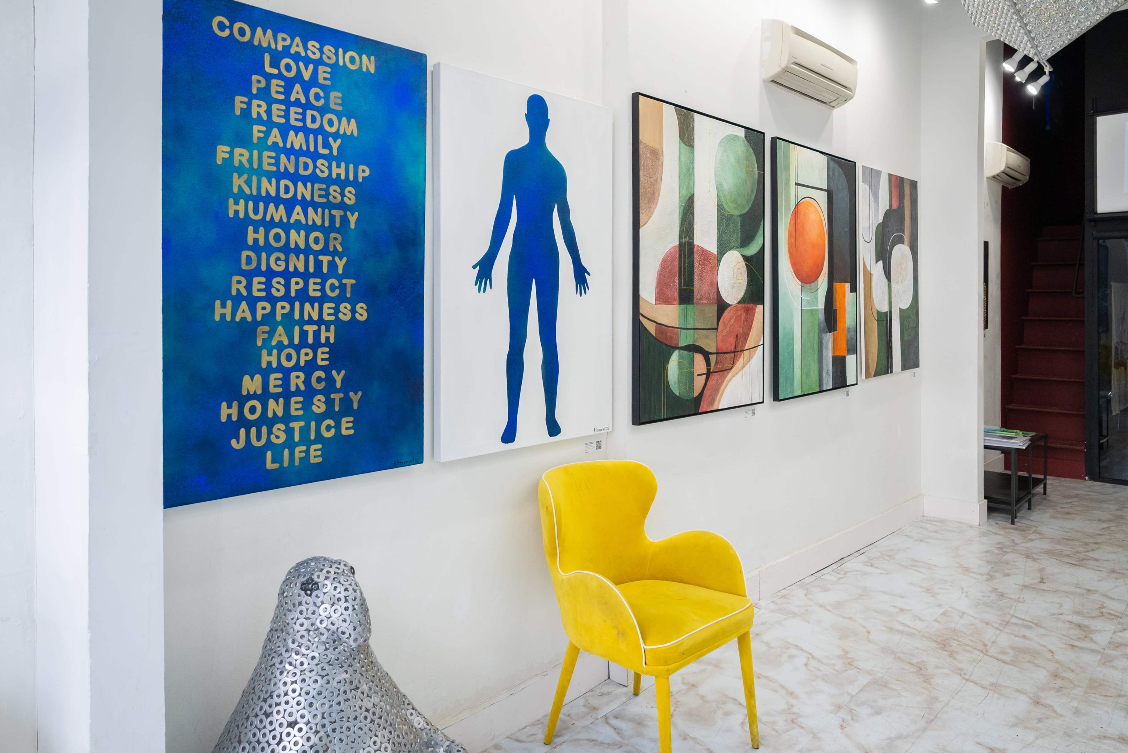 A gallery interior features vibrant wall art with various themes, alongside a yellow accent chair and a modern sculpture.