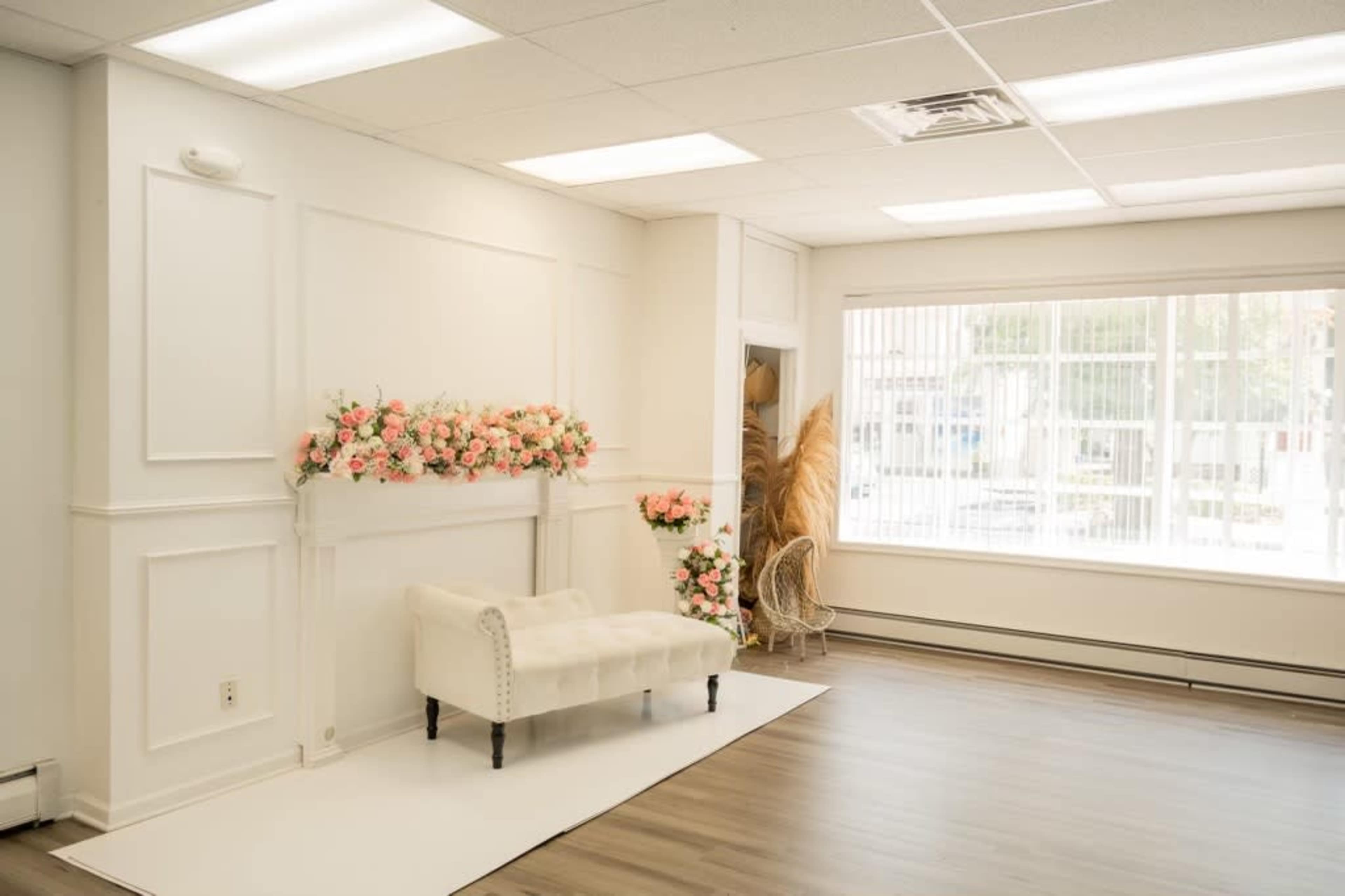A bright, minimalist room with a white wall featuring floral decorations, a white bench, and large windows allowing natural light.