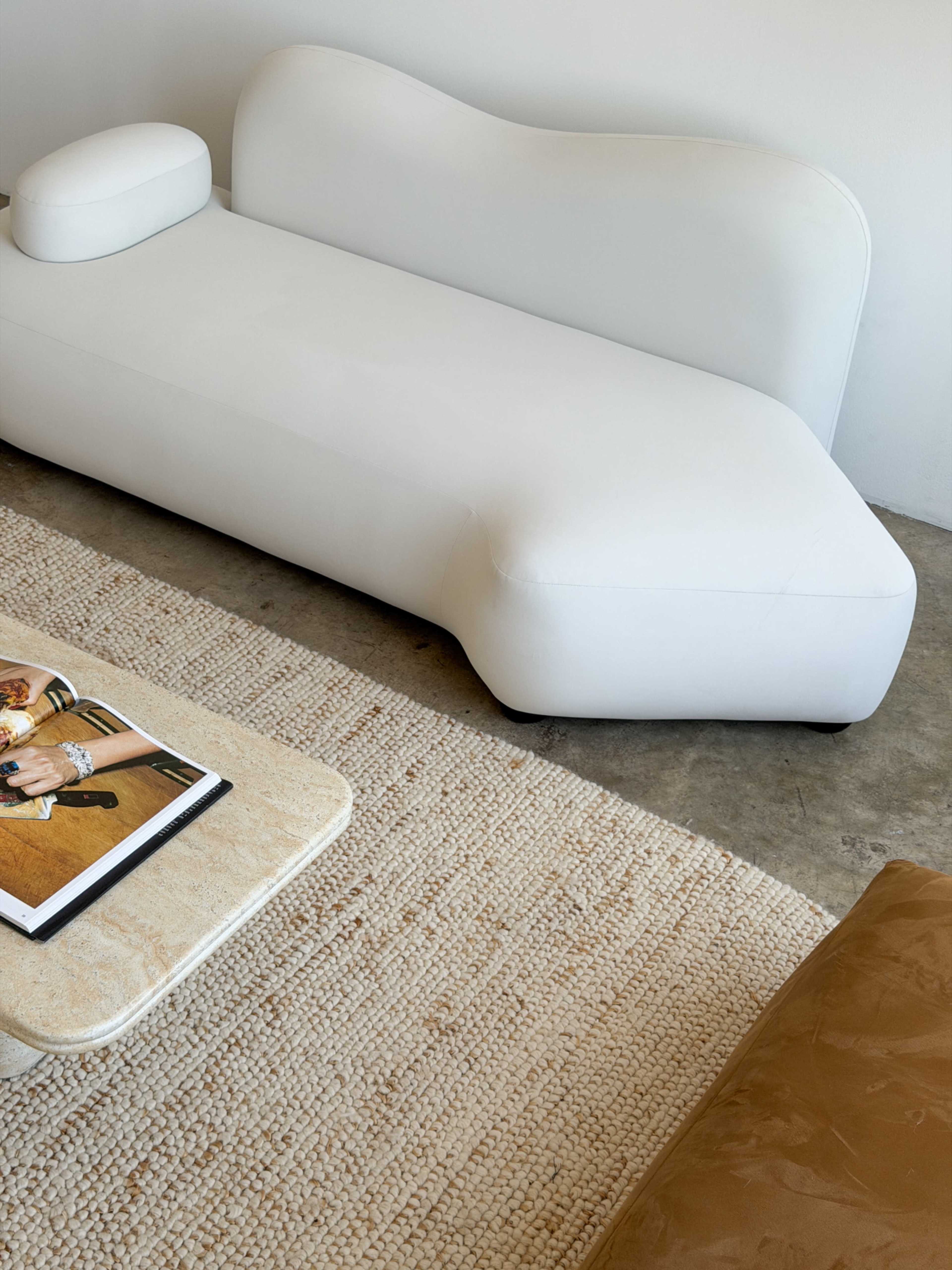 The image shows a modern, white, curved sofa positioned next to a textured rug and a coffee table with a magazine on it.