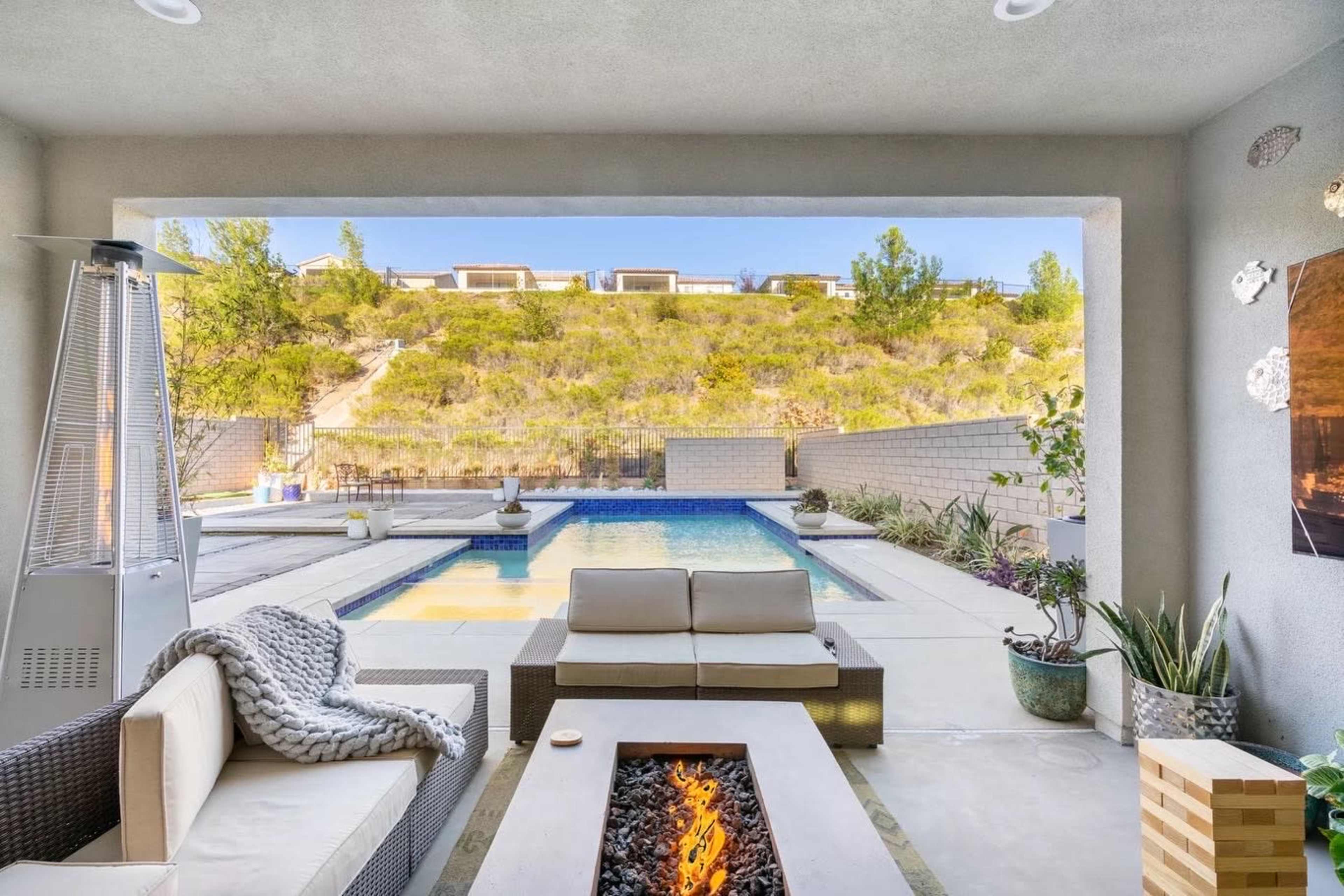A modern outdoor lounge area features a fire pit and seating beside a pool with a hillside backdrop.