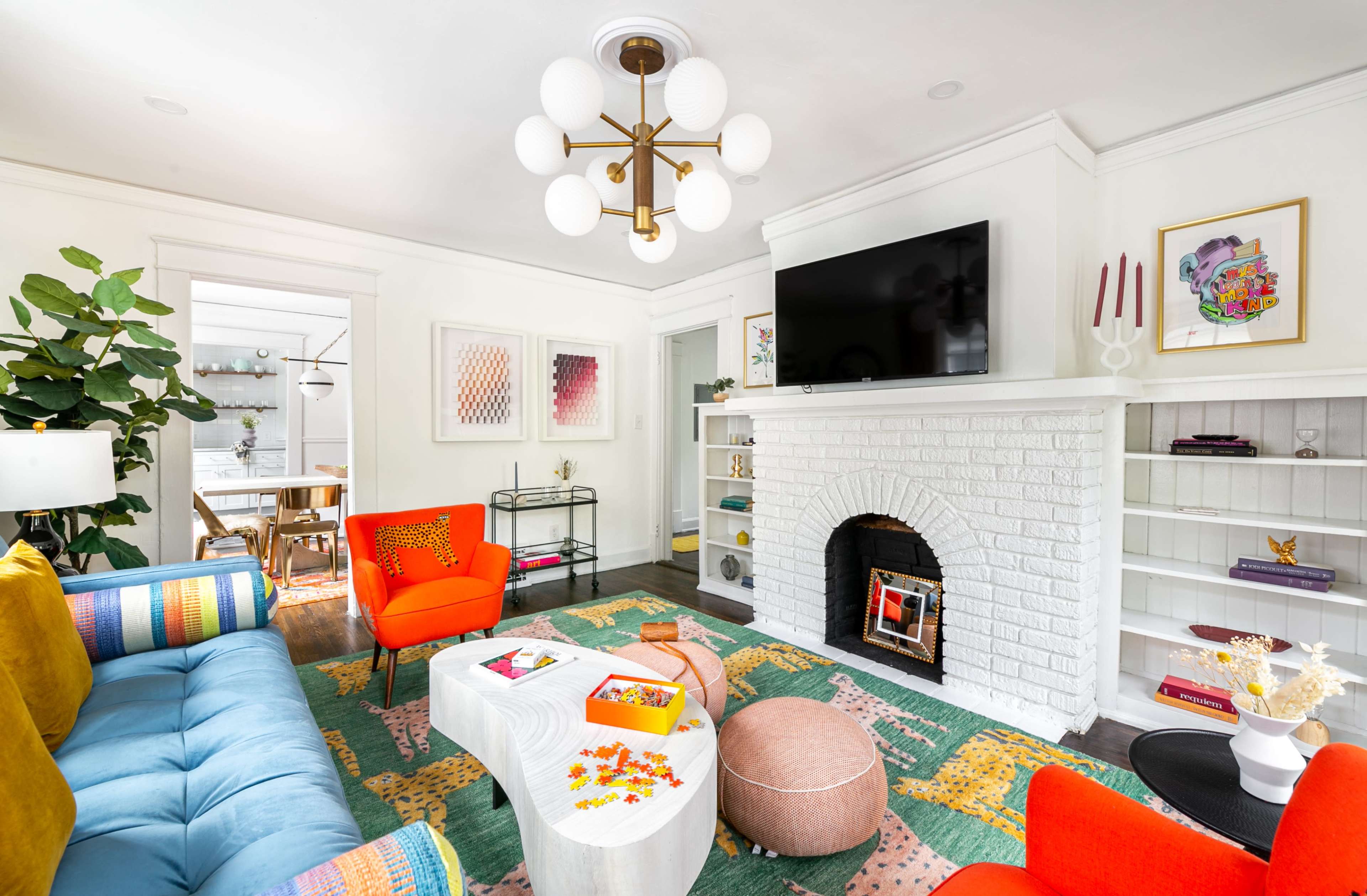 A living room features a colorful sofa, bright chairs, a white brick fireplace with a mounted television, and a coffee table surrounded by decorative items and plants.