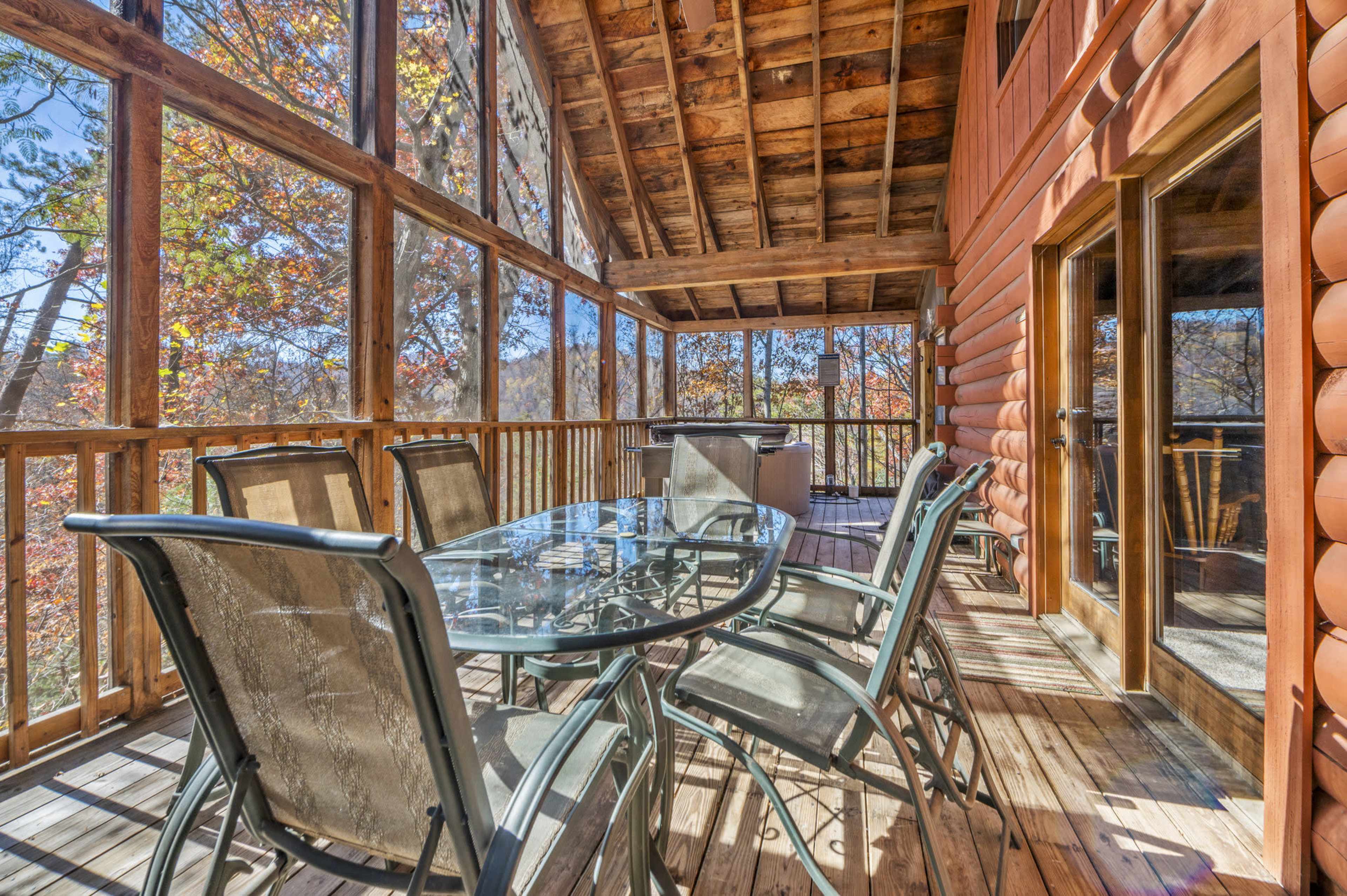 A screened porch features a glass-top dining table surrounded by metal chairs, overlooking a wooded landscape with autumn foliage.