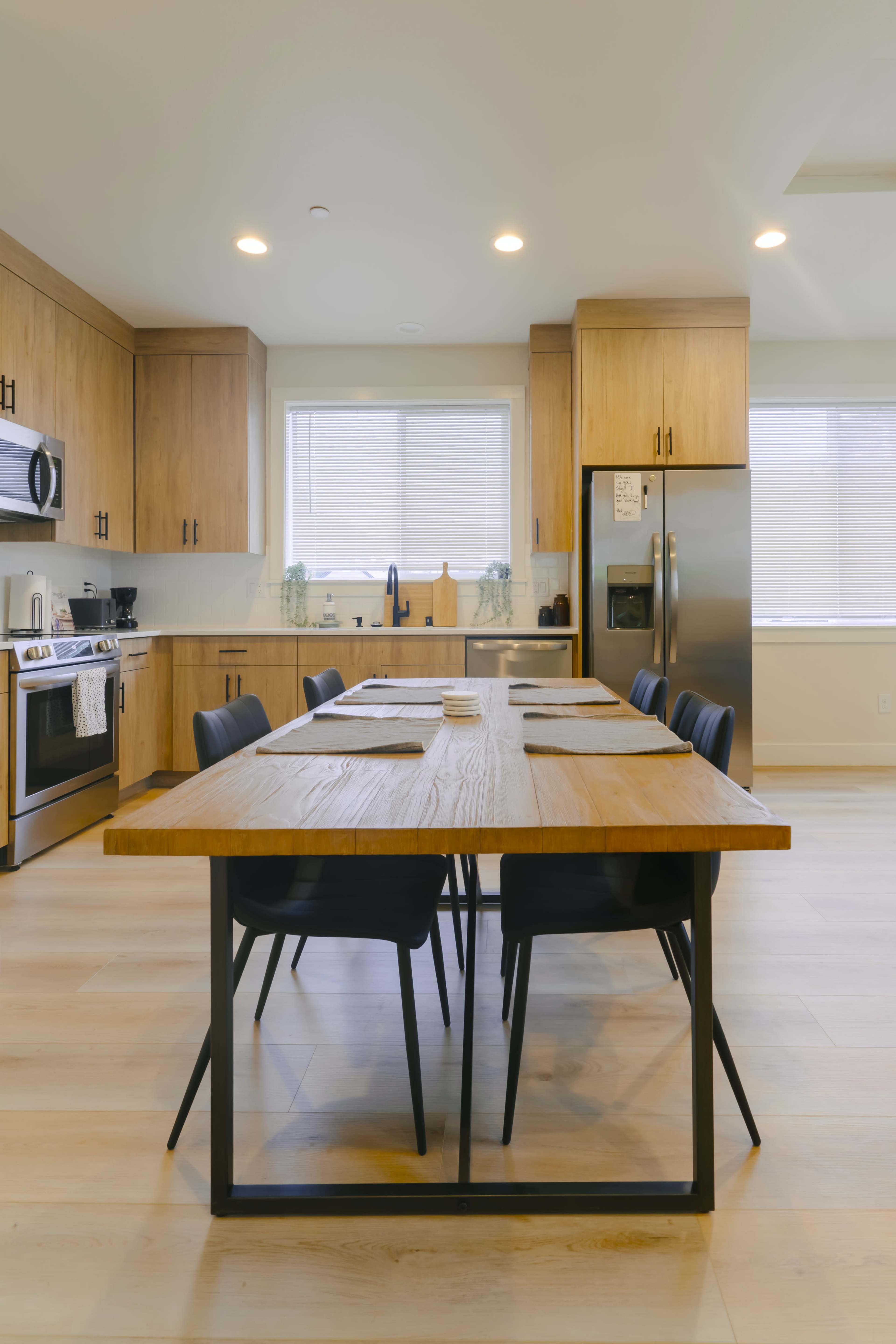 A modern kitchen features wooden cabinetry, stainless steel appliances, and a large dining table with black chairs.