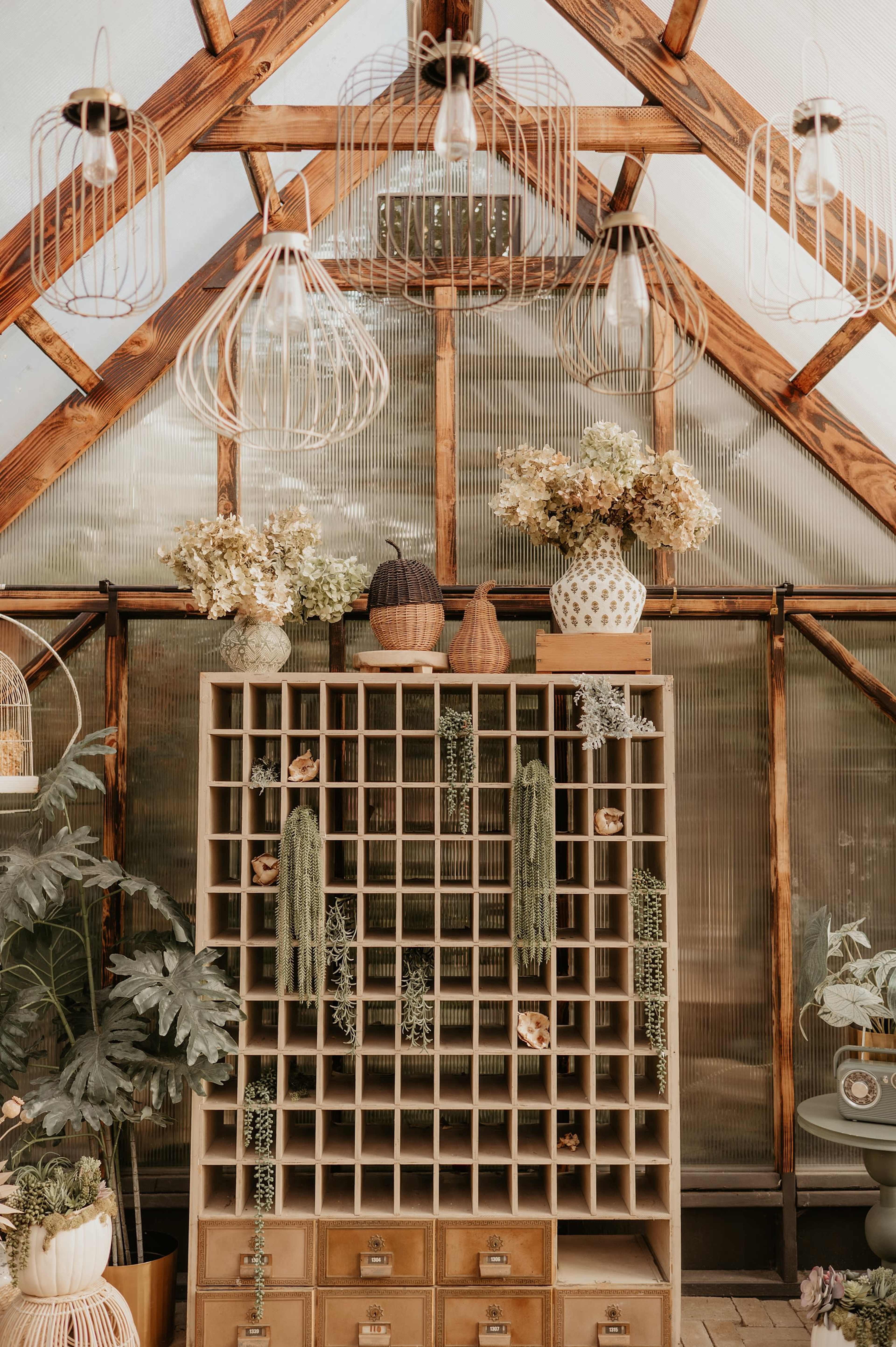 The image shows a wooden display unit filled with decorative items and plants inside a greenhouse, with hanging light fixtures above.