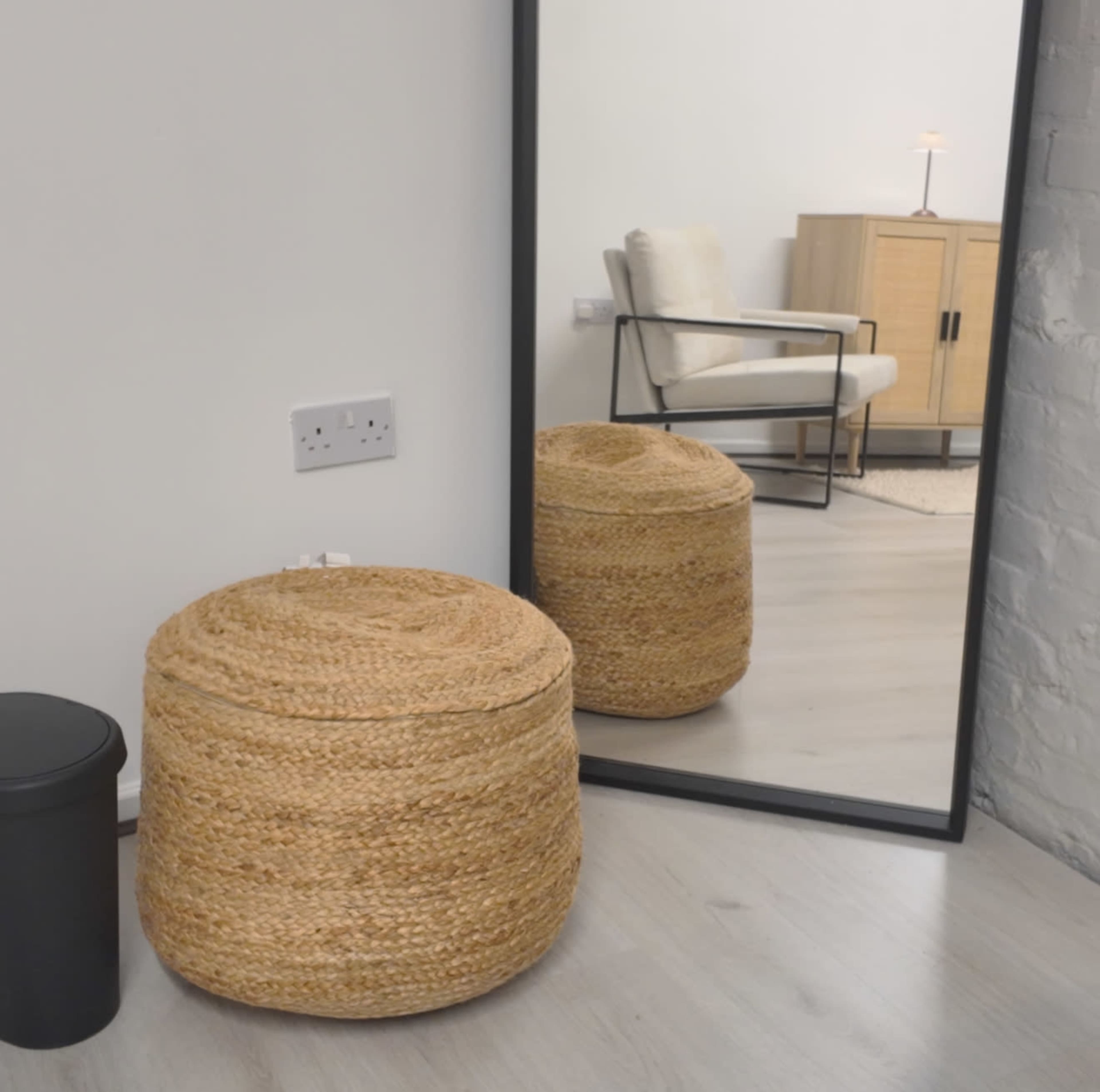 The image shows a woven round ottoman next to a black cylindrical trash can, with a large mirror reflecting a chair and a cabinet in a minimalist room.