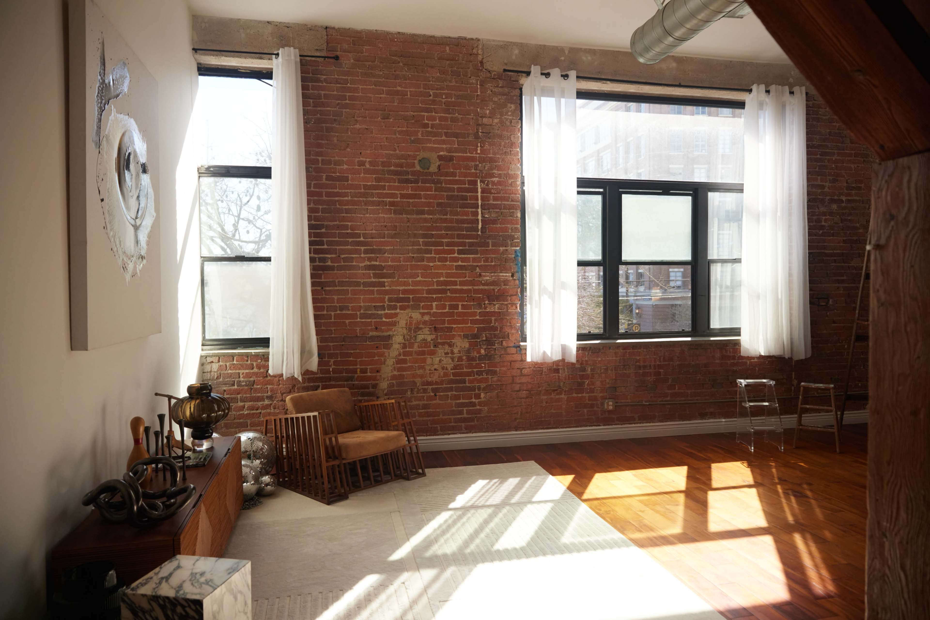 The image shows a spacious room with exposed brick walls, large windows, wooden flooring, and minimalist furniture.