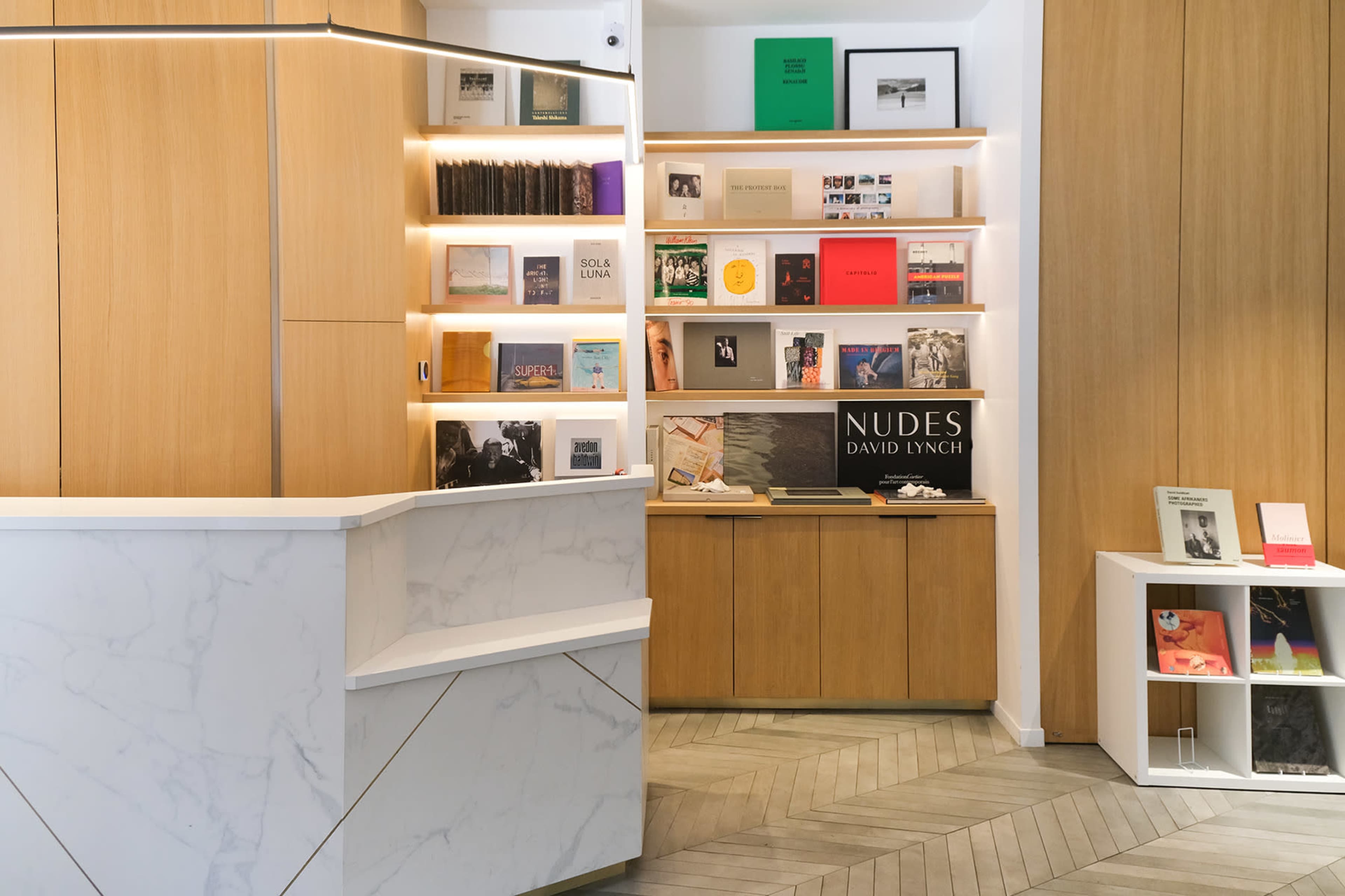 The image shows a modern interior space featuring a minimalist reception desk and wooden shelves displaying various books and art publications.