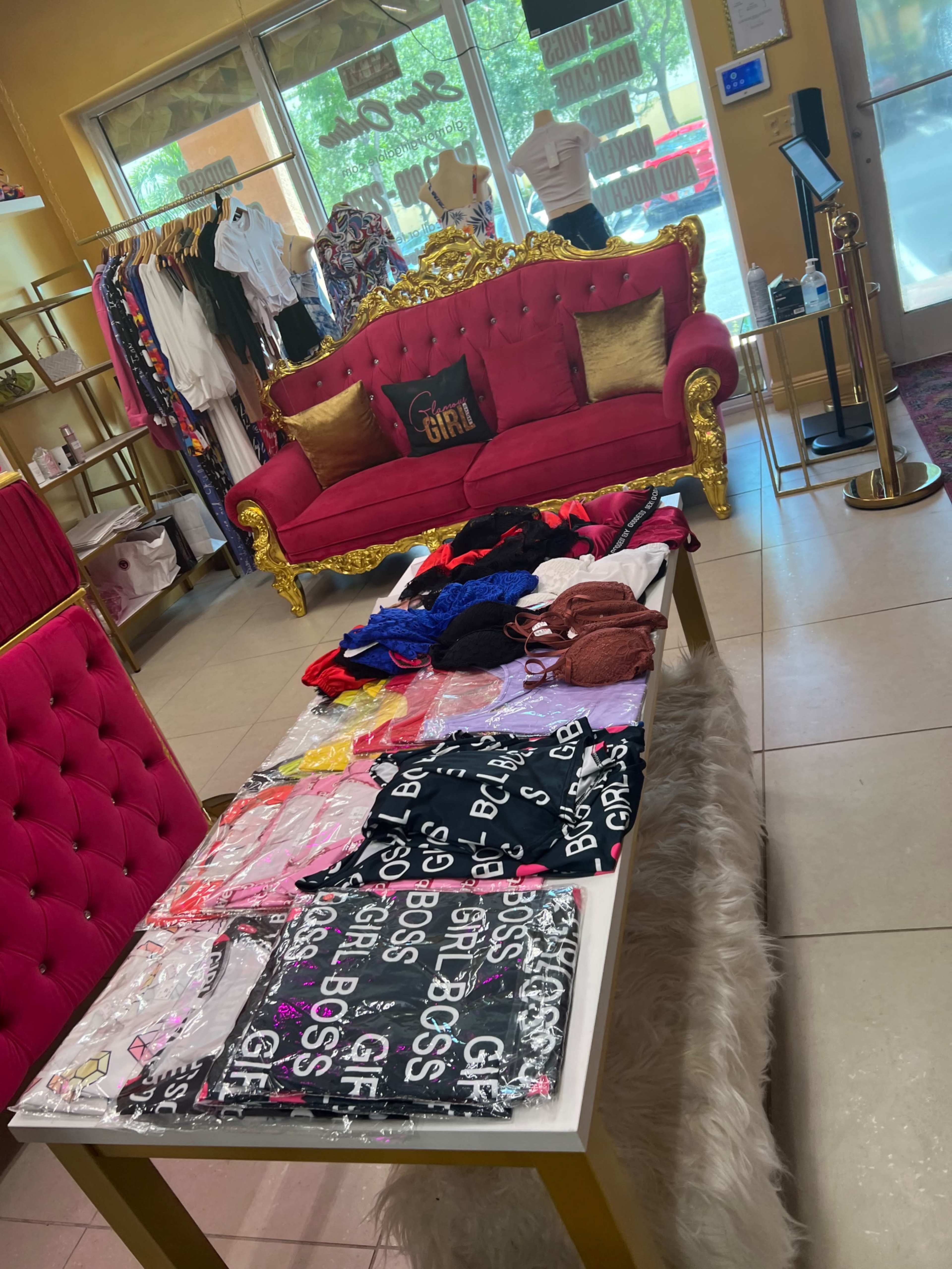 The image shows a boutique interior featuring a pink sofa, a clothing display table with various apparel items, and a rack of hanging garments in the background.