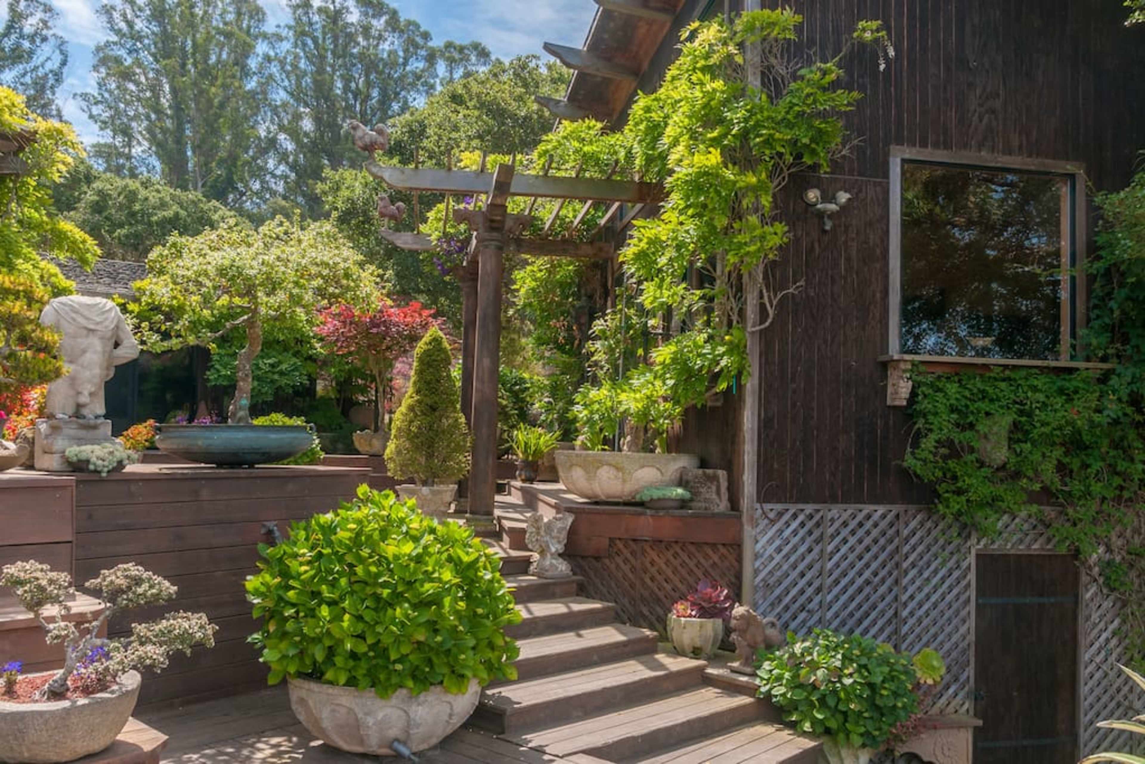 A landscaped garden with various plants, stone sculptures, and a wooden staircase leading to a cozy wooden structure.