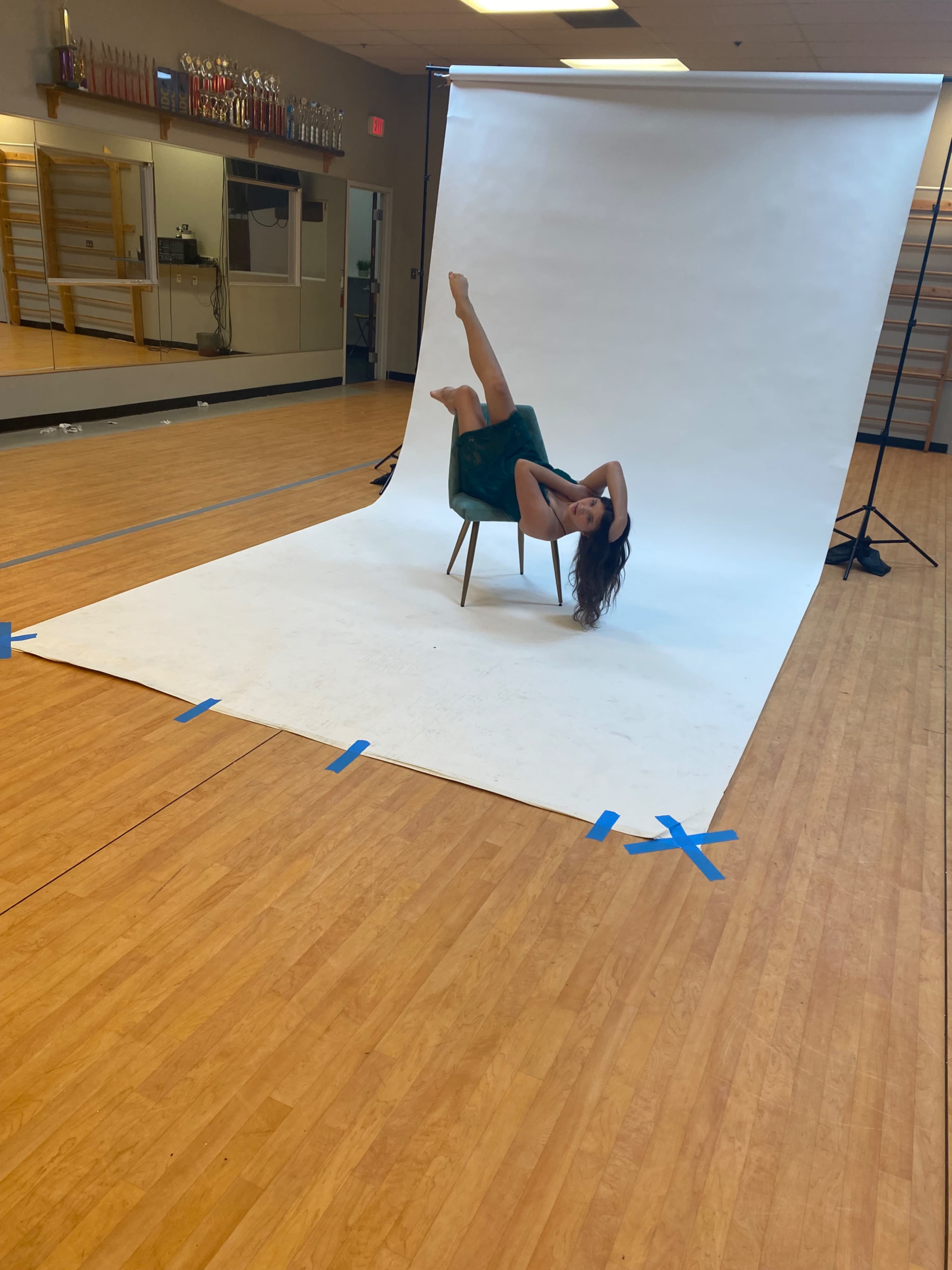 A person posed on a chair, with one leg raised, against a plain white backdrop in a well-lit studio space.