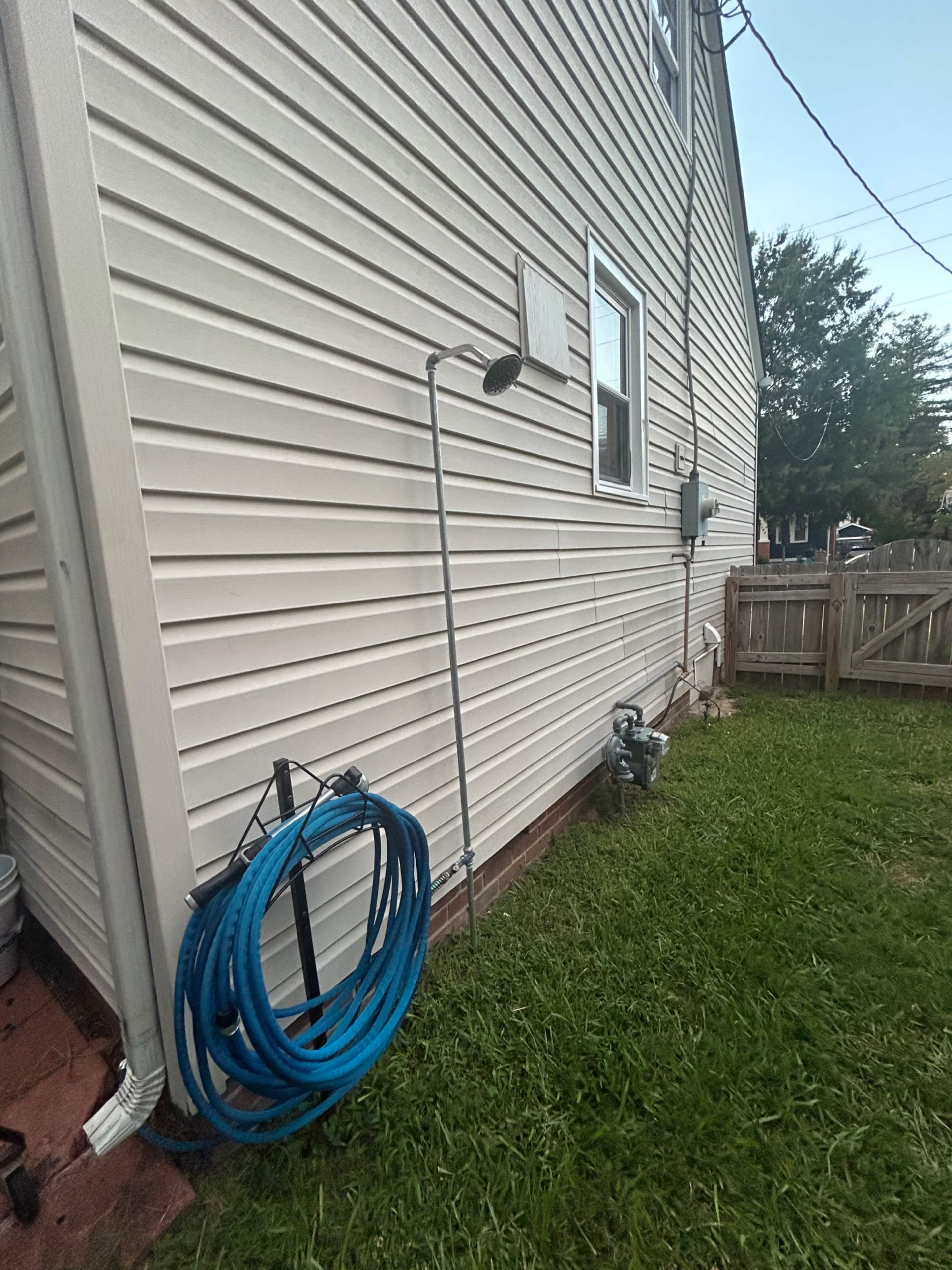 A plastic garden hose coiled on the ground is positioned next to an outdoor shower fixture attached to the side of a house, with a grassy area nearby.