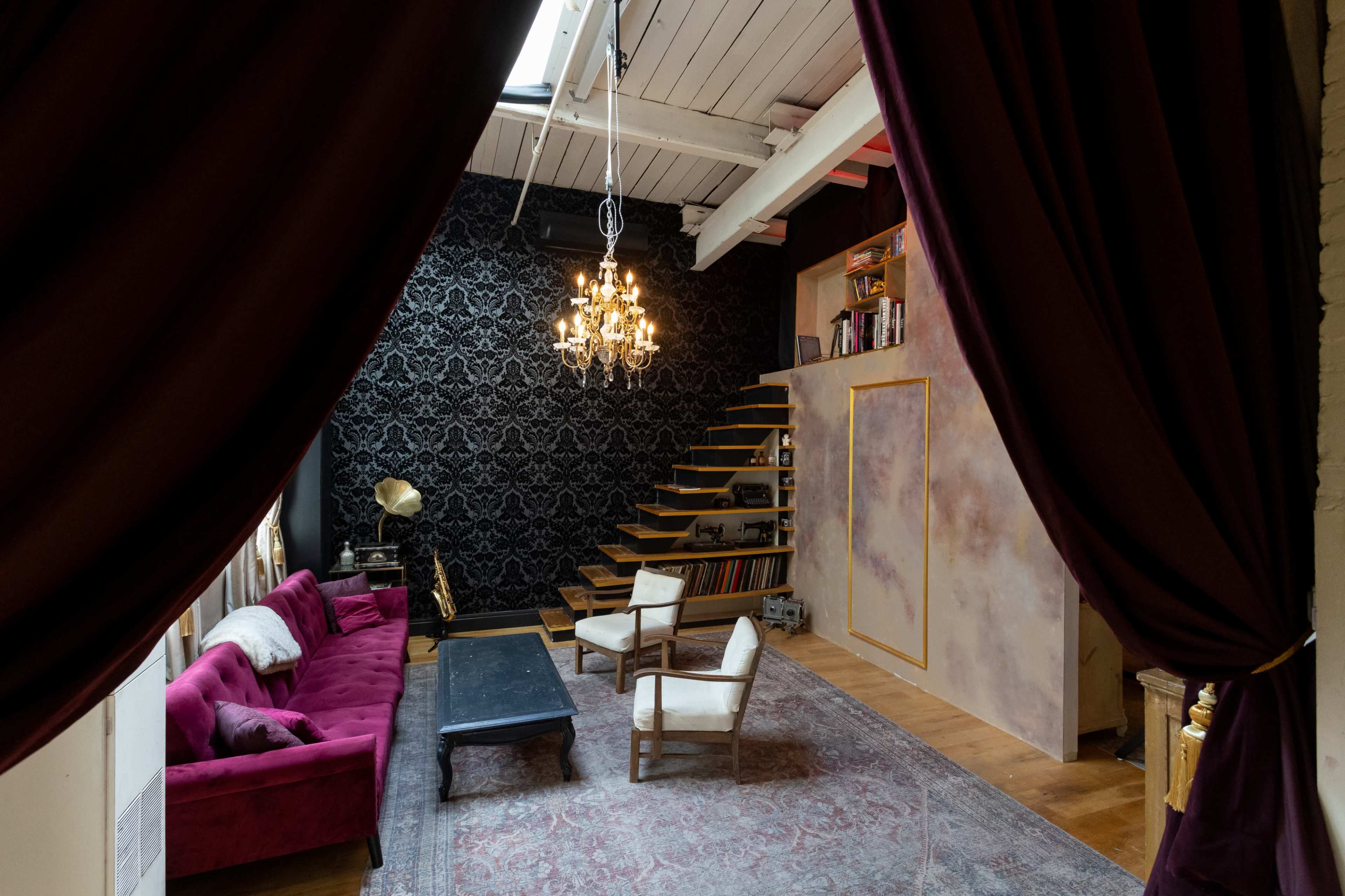 A spacious living area features a velvet couch, two armchairs, a dark coffee table, and a staircase lined with bookshelves against a patterned wall, all framed by burgundy curtains.