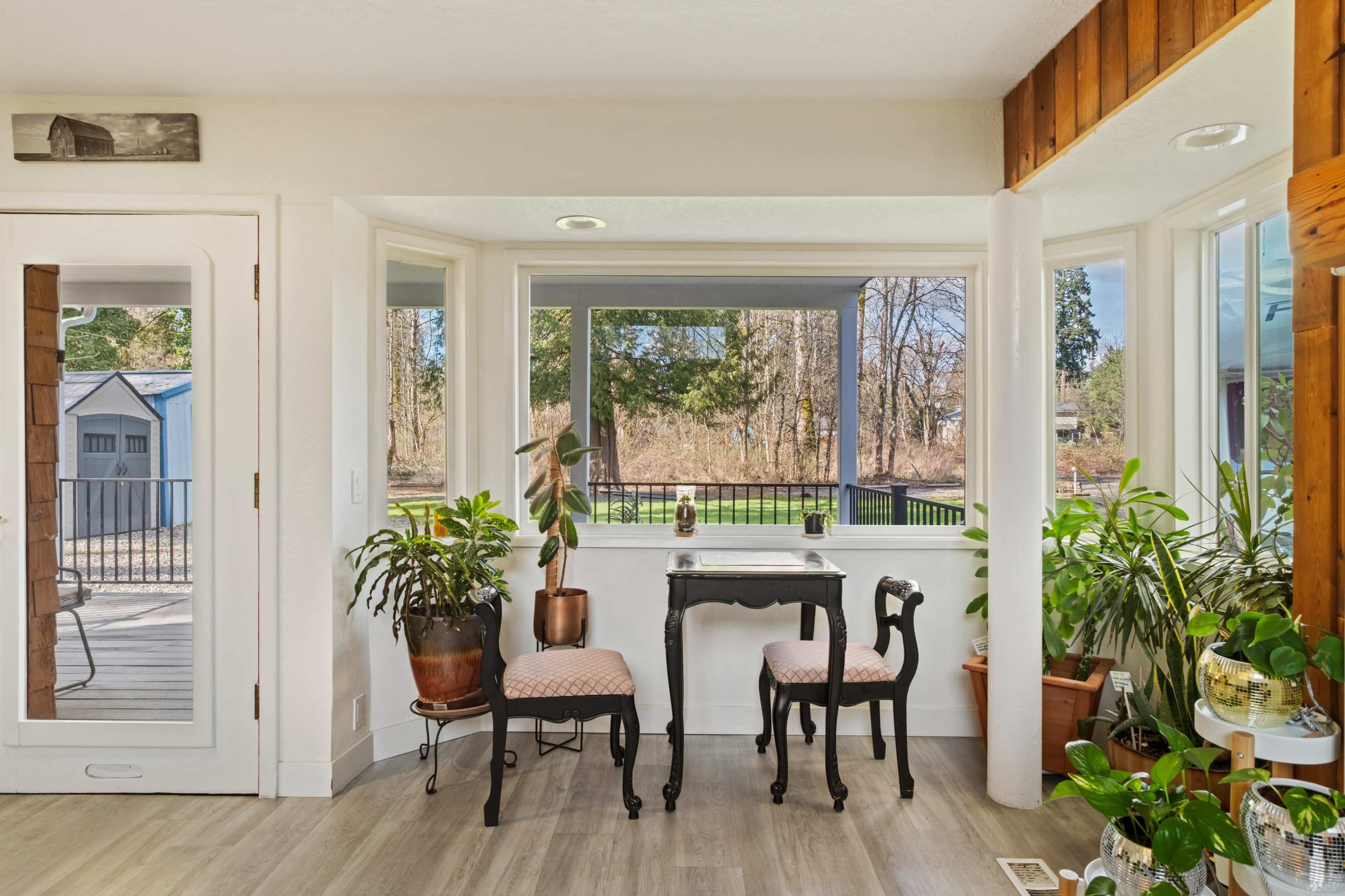 The image shows a bright sunroom with two chairs and a small table, surrounded by potted plants and large windows overlooking a natural landscape.