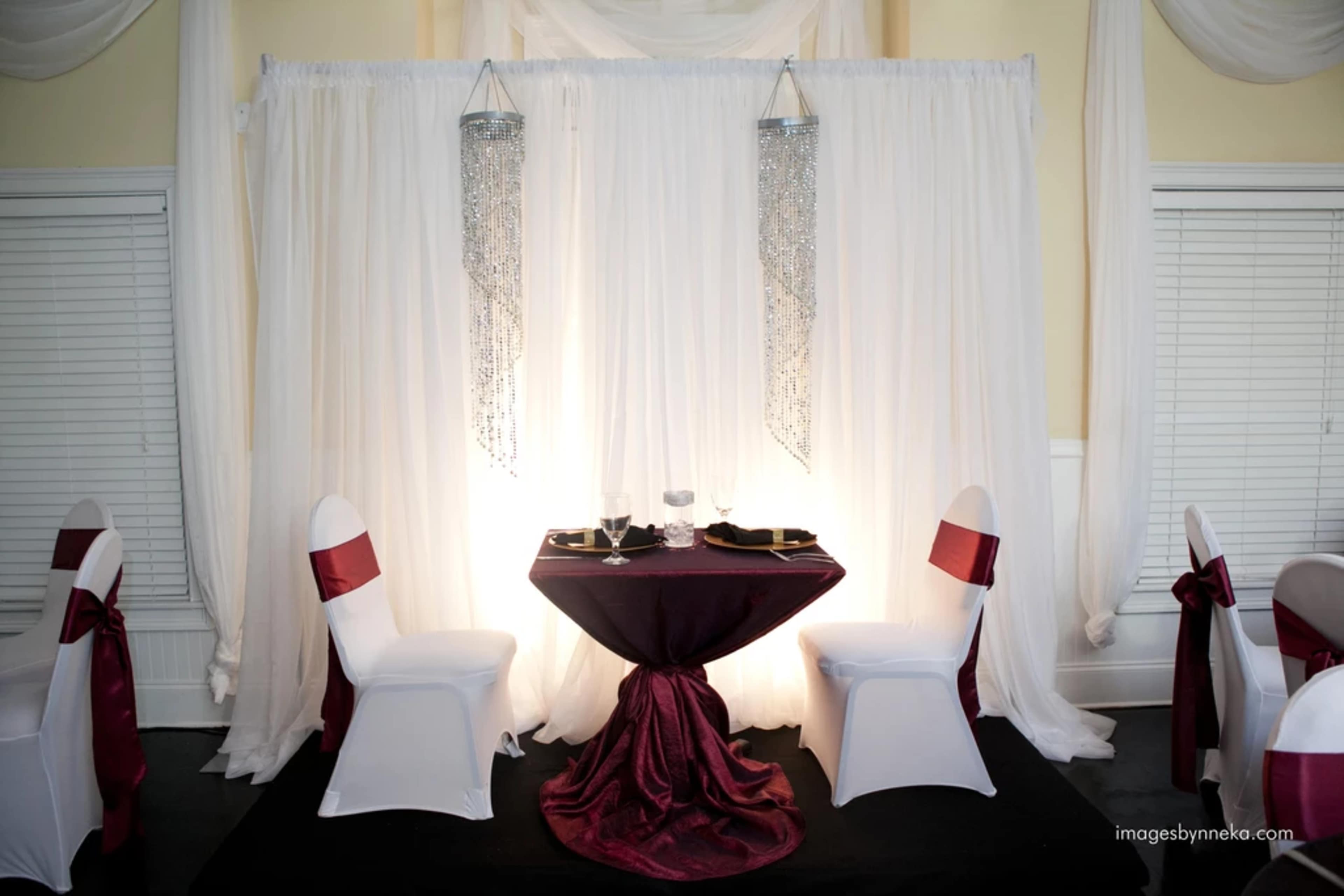 A small round table with a deep red tablecloth is set for two, surrounded by chairs draped in white with red sashes, in front of a backdrop of flowing white drapery and hanging metallic accents.