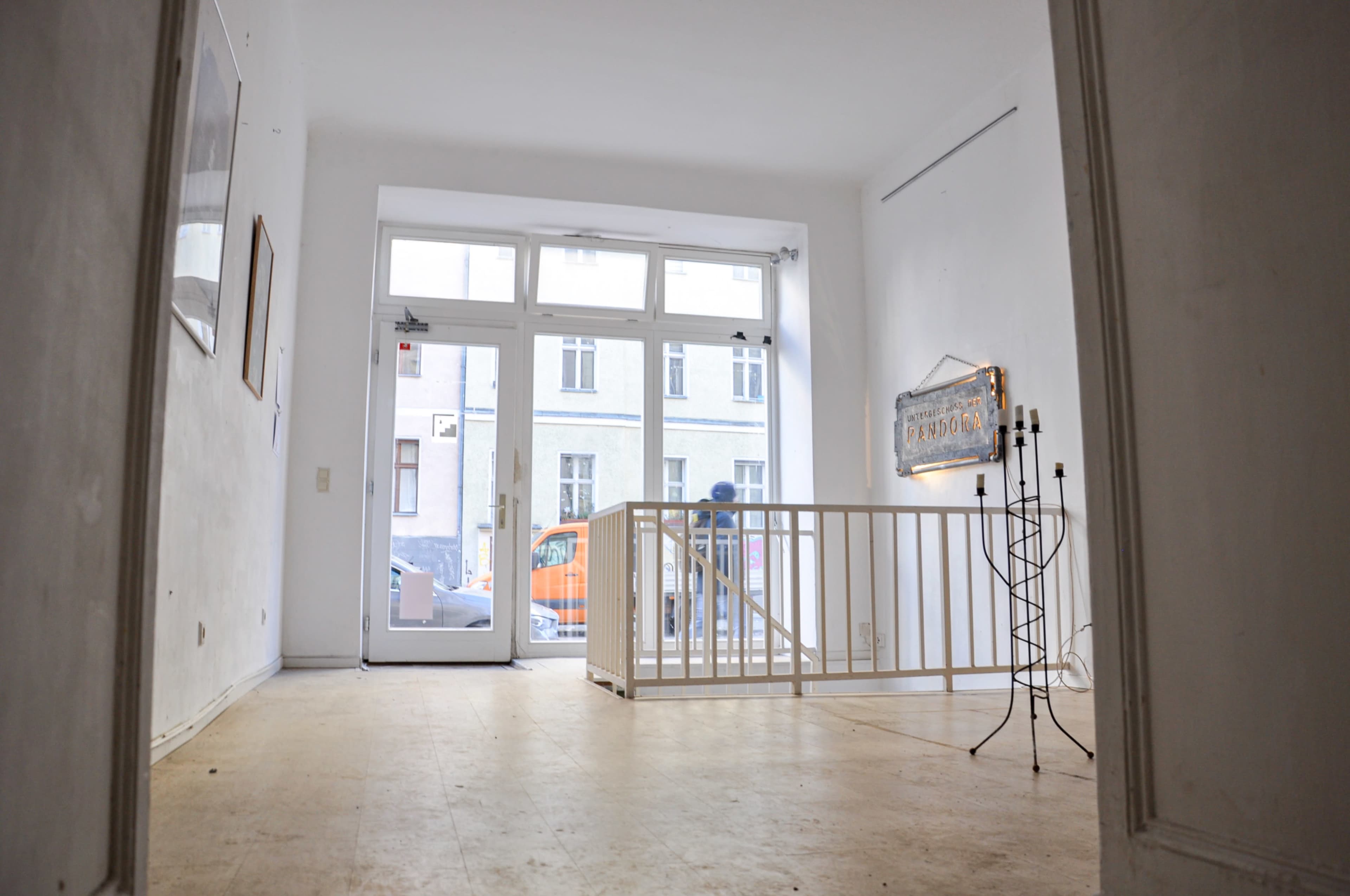 The image shows a spacious, empty room with white walls and wooden flooring, featuring a railing near the entrance and a decorative light fixture on the wall.