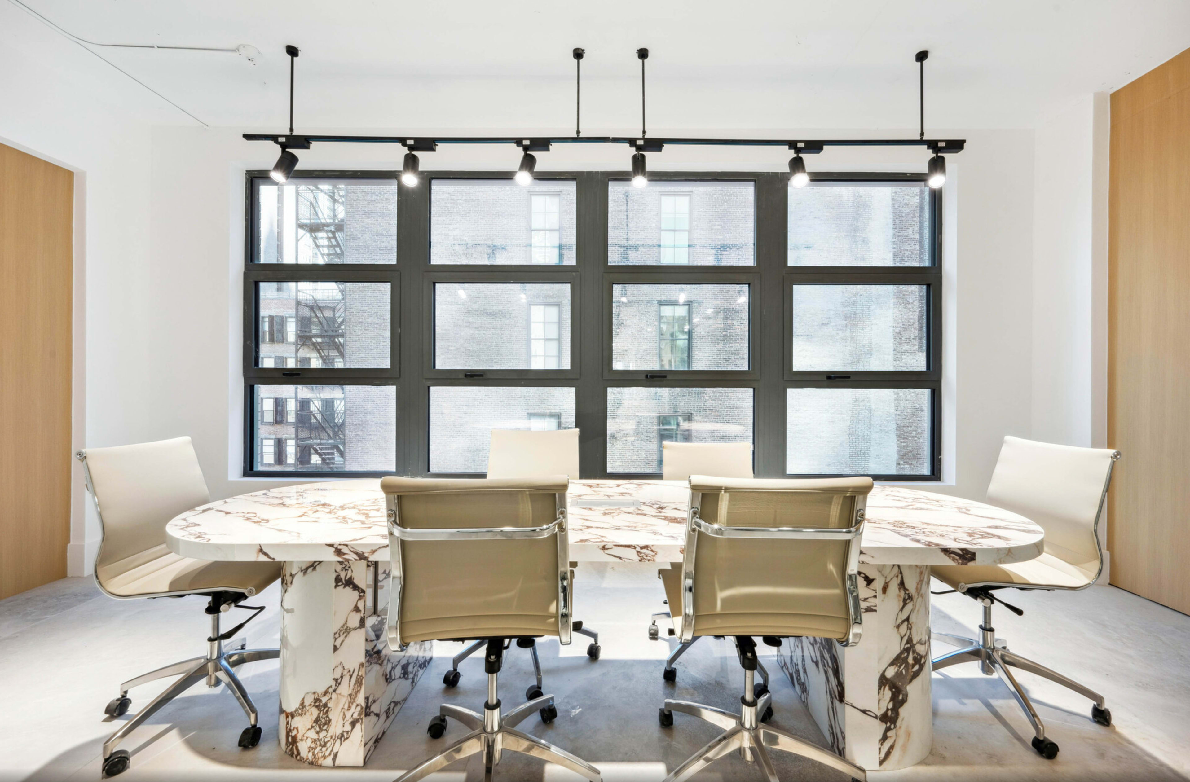 A modern conference room features a round table with a marble design and six white swivel chairs, illuminated by overhead track lighting.