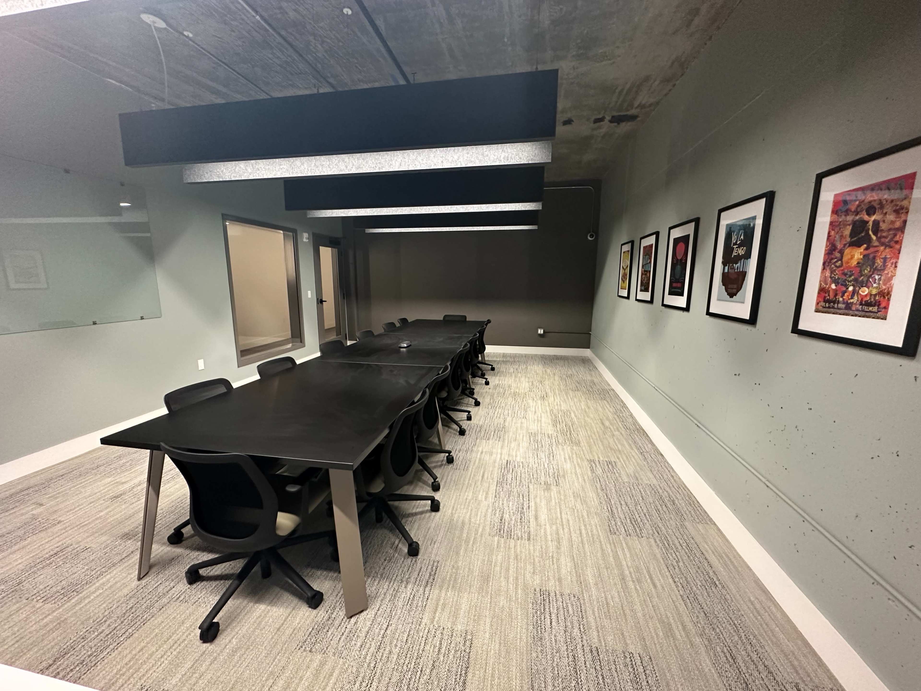 A long conference table with rolling chairs is positioned in a modern meeting room featuring framed artwork on the walls.