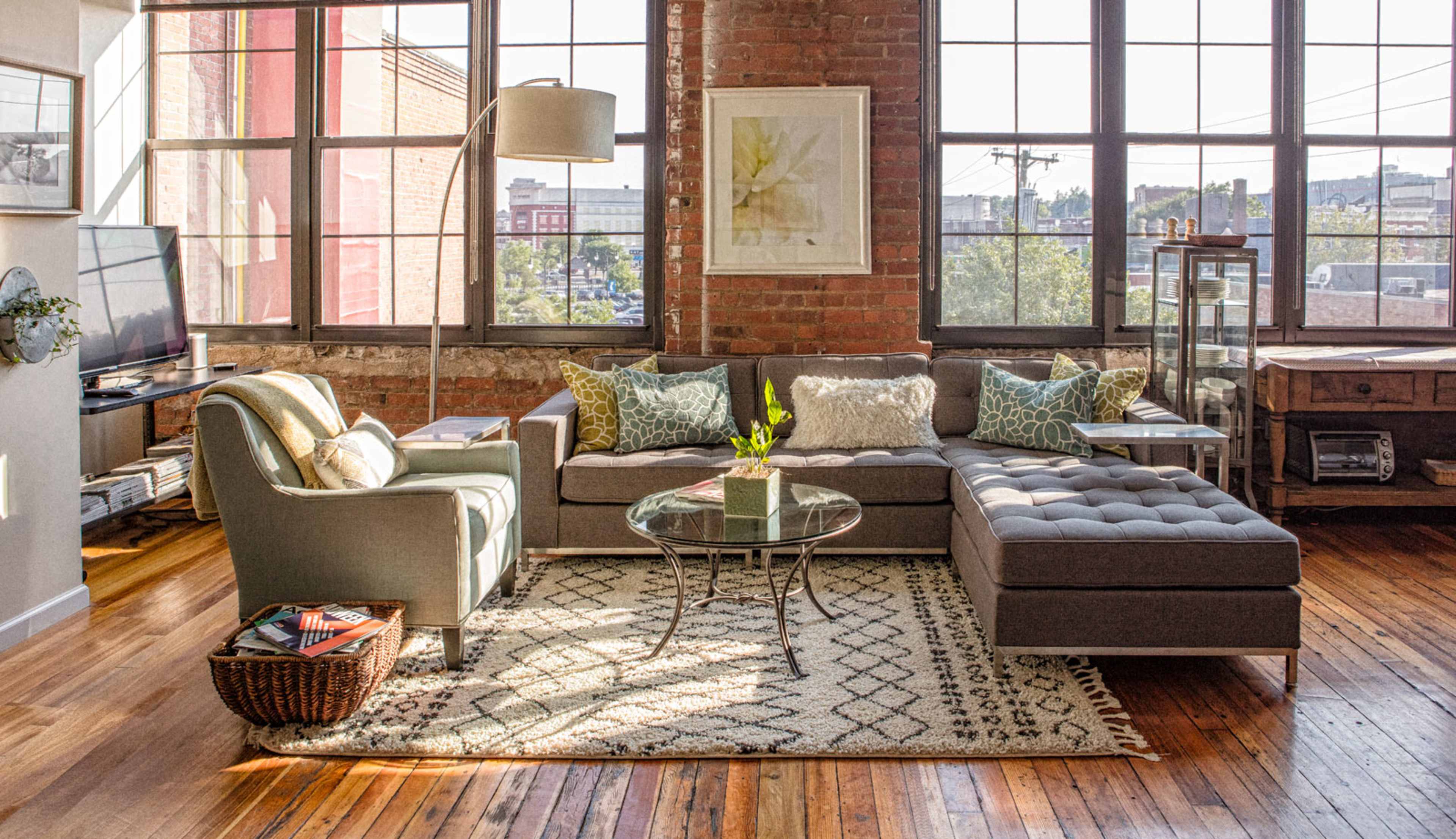 A cozy living room with a sectional sofa, a glass coffee table, and large windows allowing natural light to fill the space.