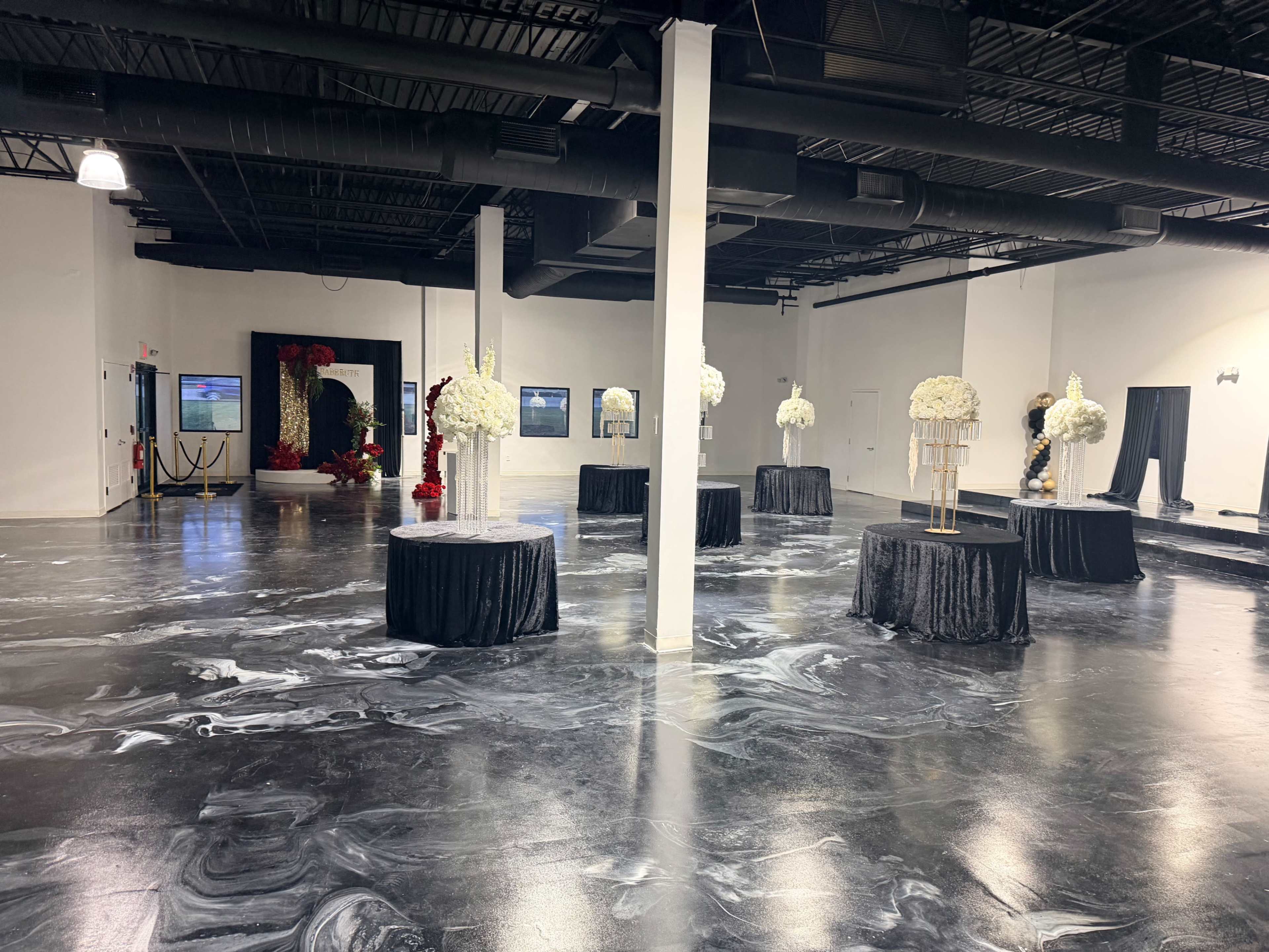 The image shows a spacious event venue with black and white marbled flooring, featuring several round tables adorned with black tablecloths and floral arrangements.