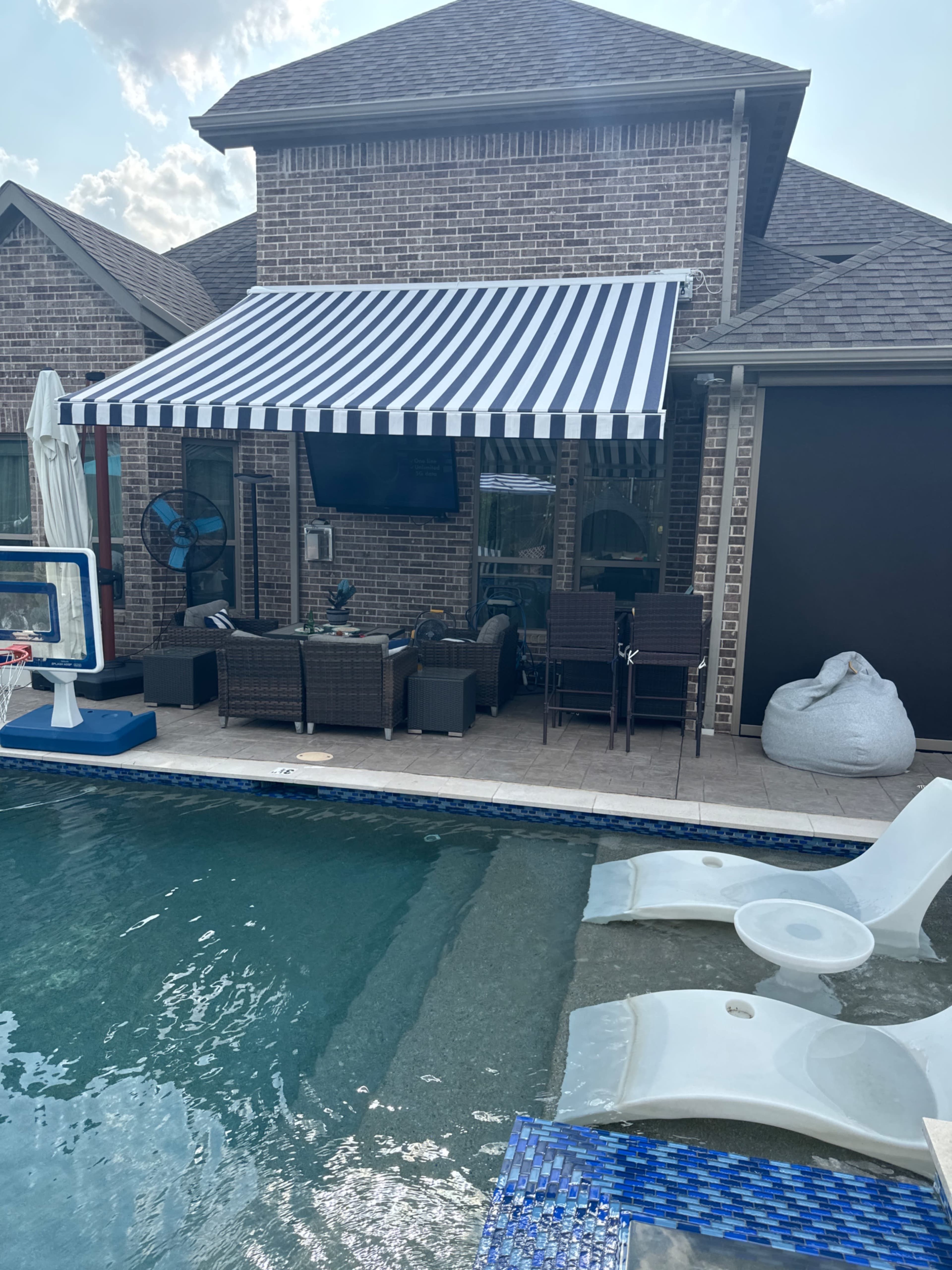 A backyard pool area with a seating arrangement under a striped awning, adjacent to a brick house.