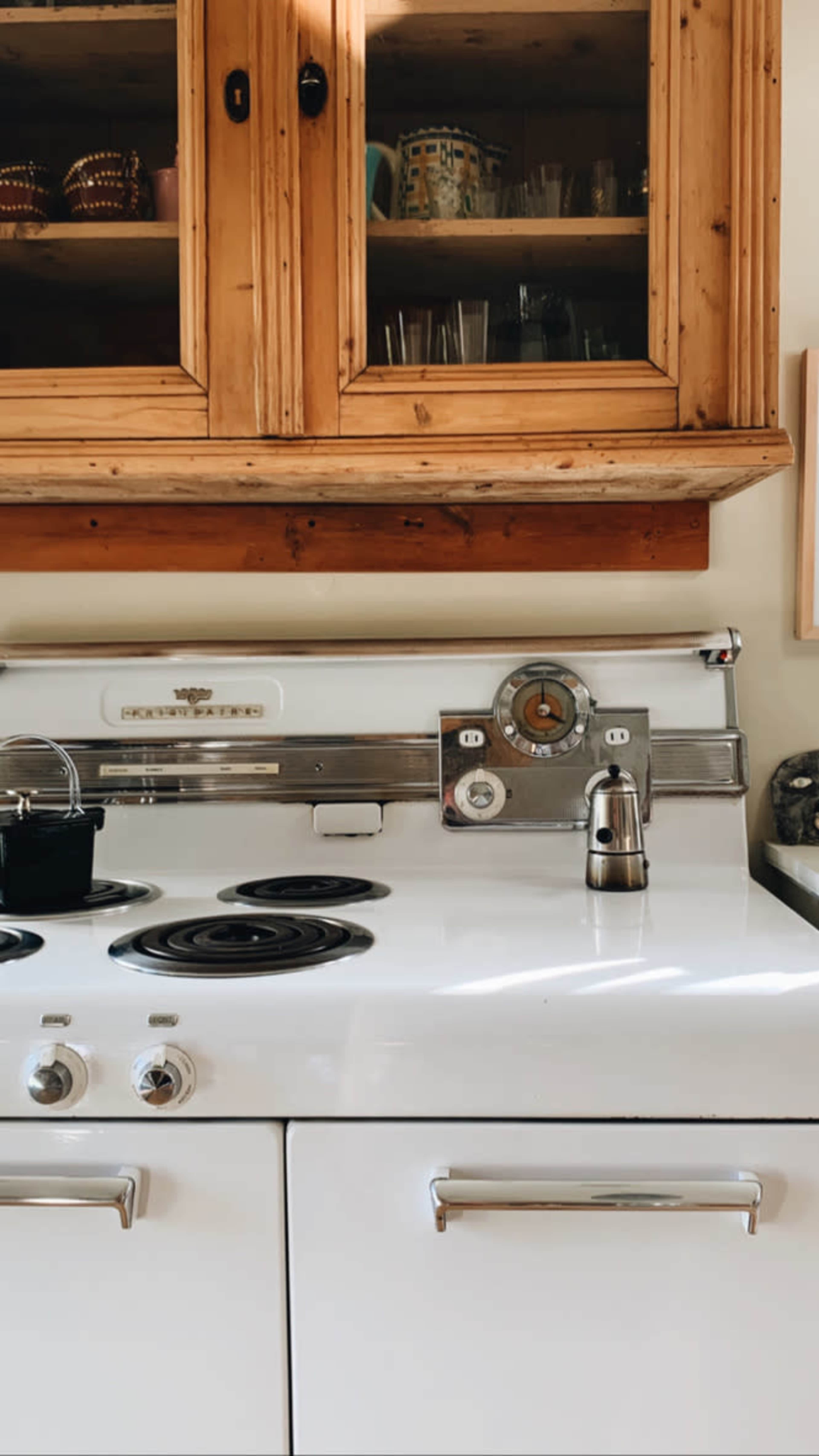 A vintage white kitchen stove with a chrome control panel and a wooden cabinet above it containing glassware.