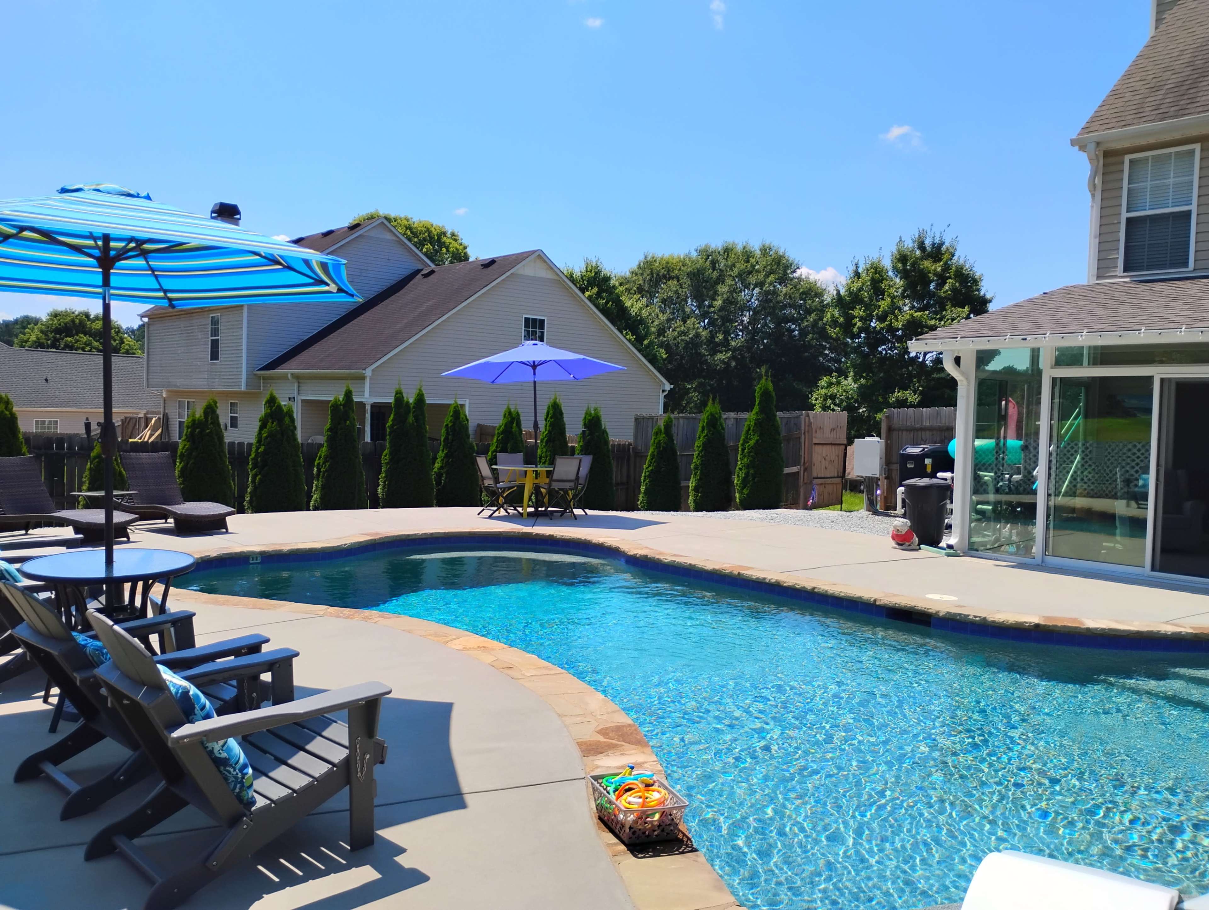 The image shows a residential backyard with a swimming pool surrounded by lounge chairs, umbrellas, and a shaded seating area.