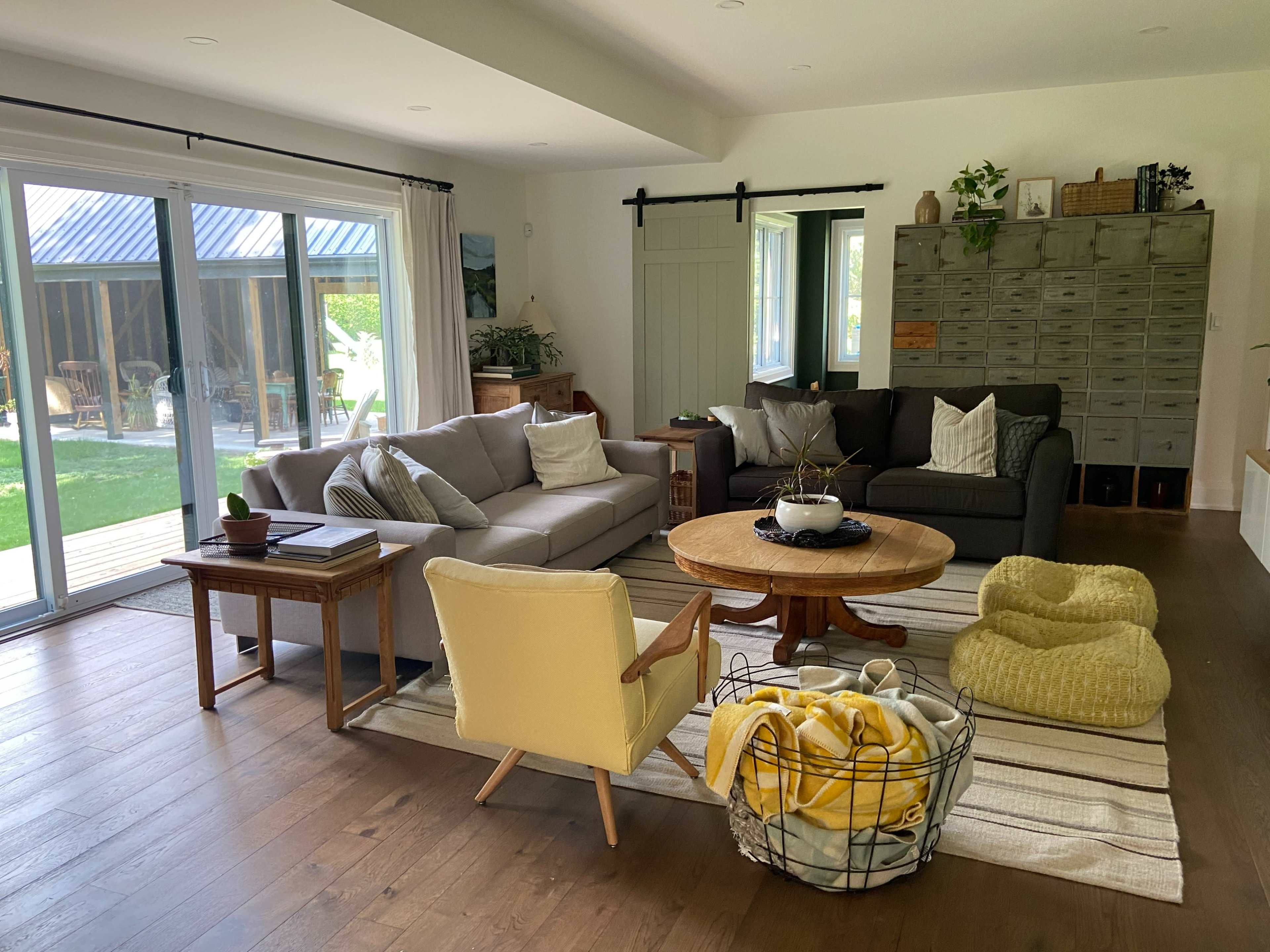 The image shows a cozy living room with a large sectional sofa, a circular coffee table, and a yellow accent chair, surrounded by natural light streaming through sliding glass doors.