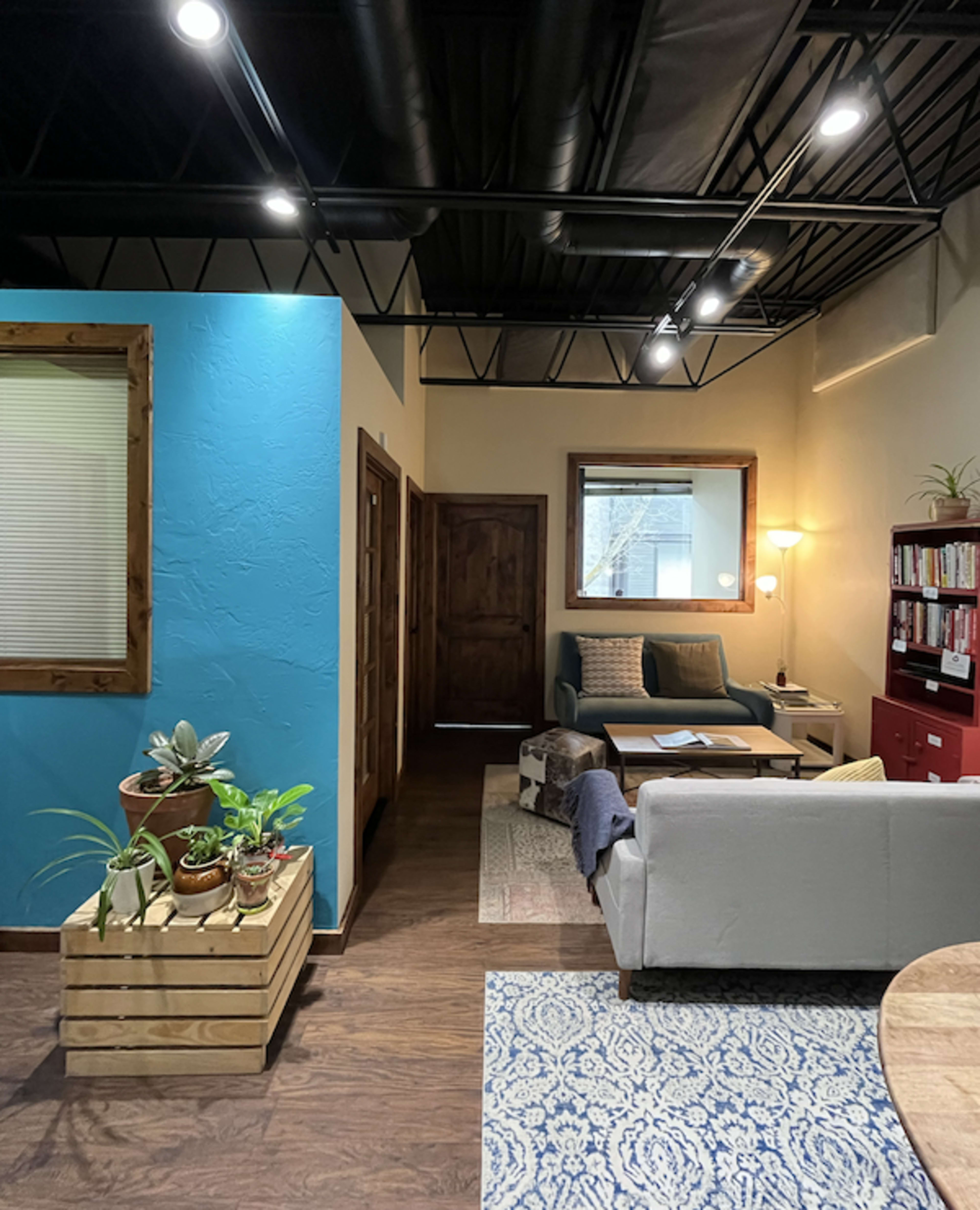 The image shows a cozy interior space with a seating area featuring a blue accent wall, a doorway leading to another room, and a wooden planter filled with plants.