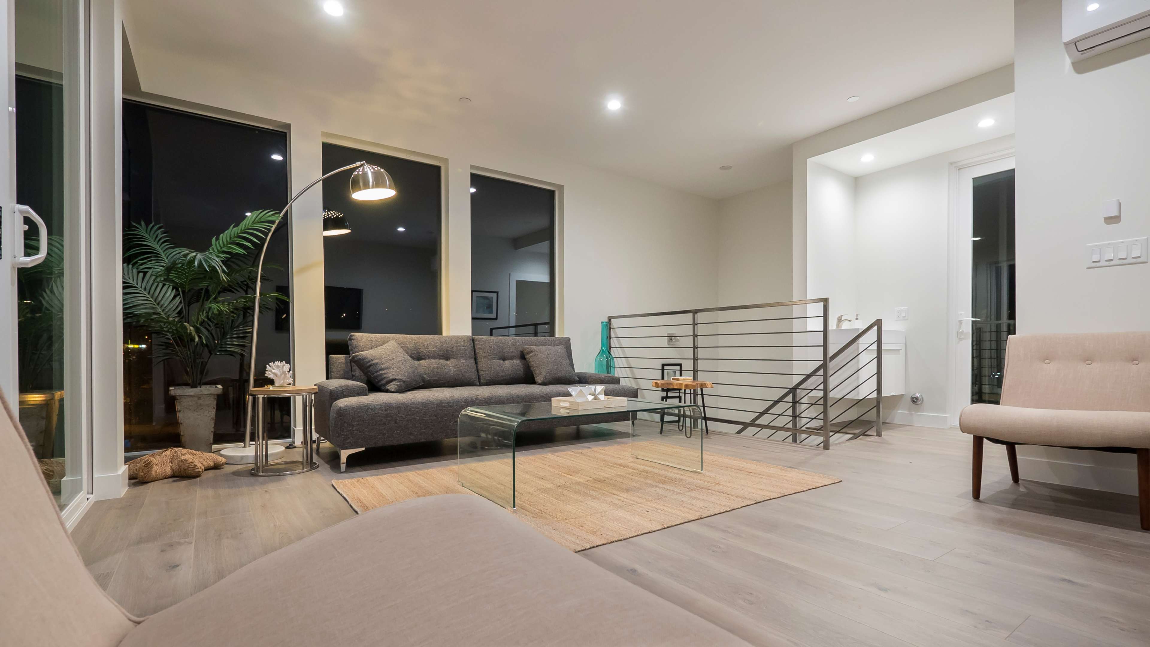 A modern living room features a gray sofa, a glass coffee table, and large windows with a view of the night sky.