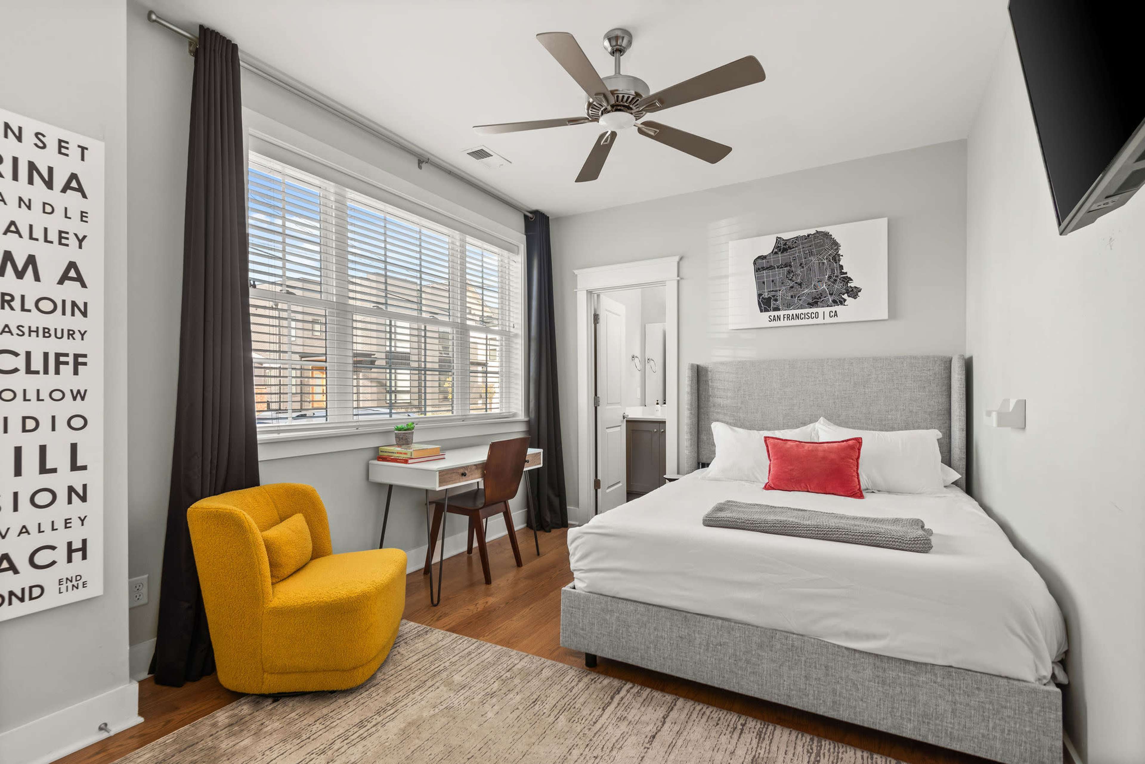 The image shows a neatly arranged bedroom featuring a bed with white linens, a yellow accent chair, a wooden desk, and a window with blinds.