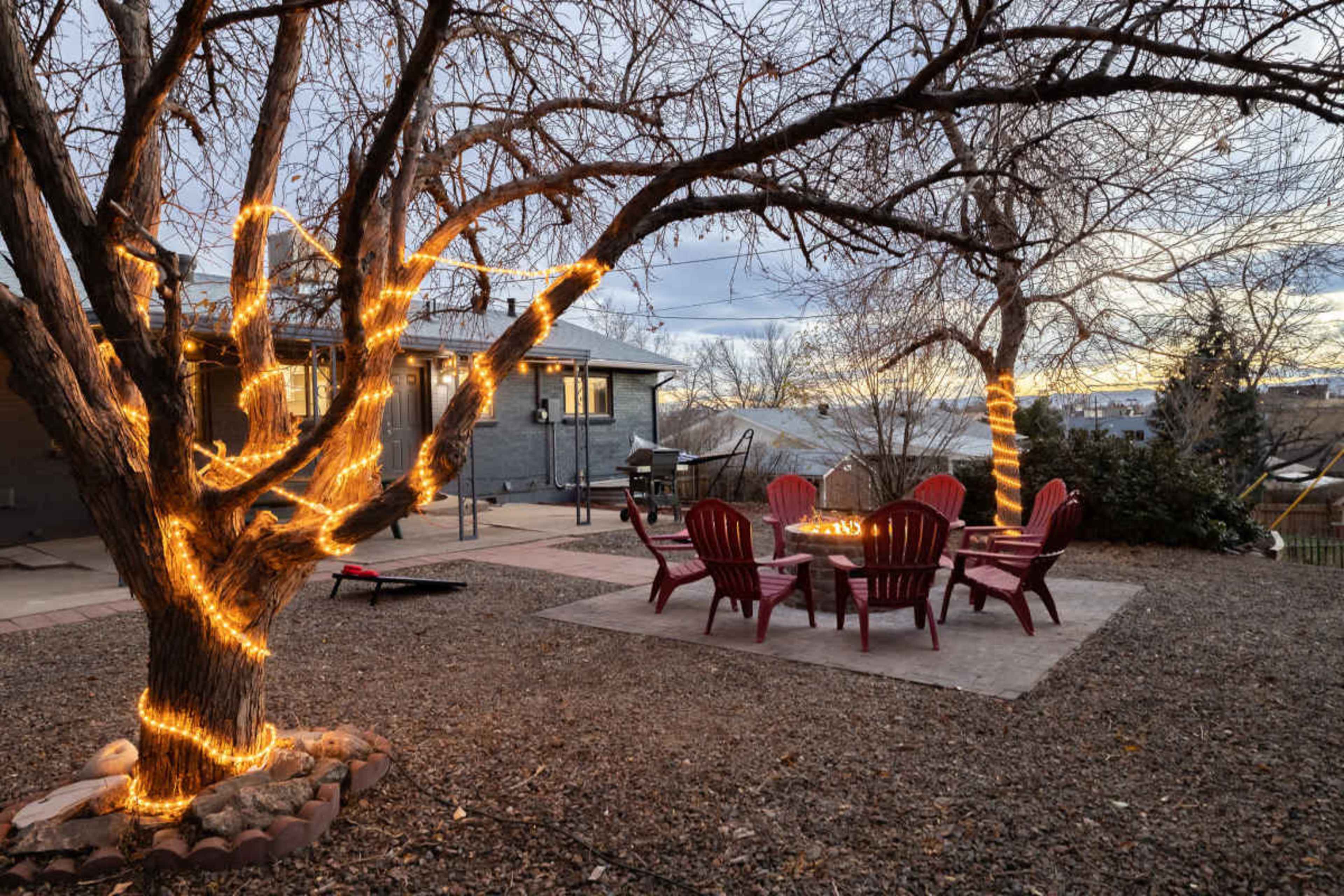 A backyard features a fire pit surrounded by red chairs and a tree wrapped in string lights during twilight.
