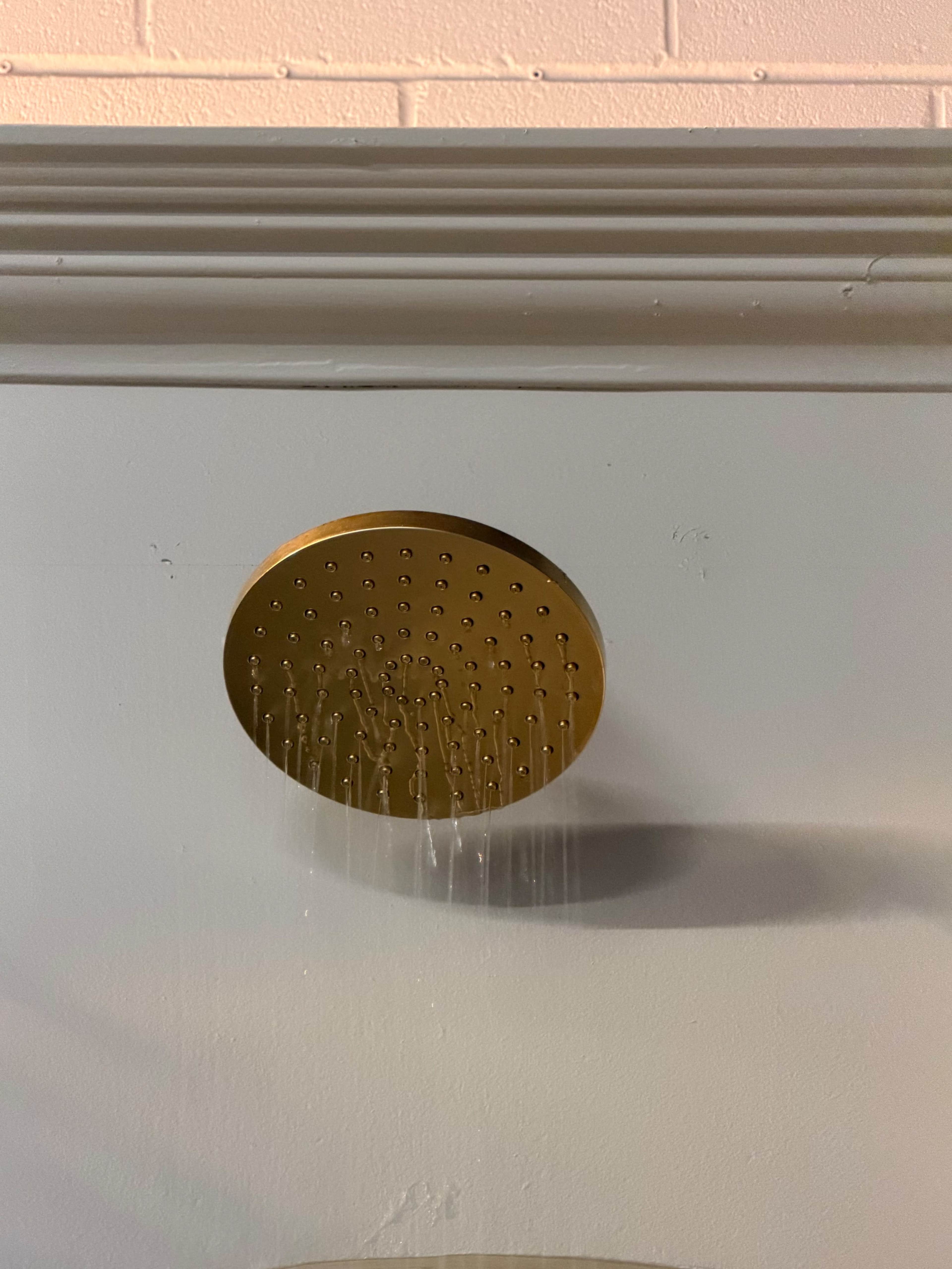 A circular gold showerhead is mounted on a white wall, with water droplets cascading downwards.