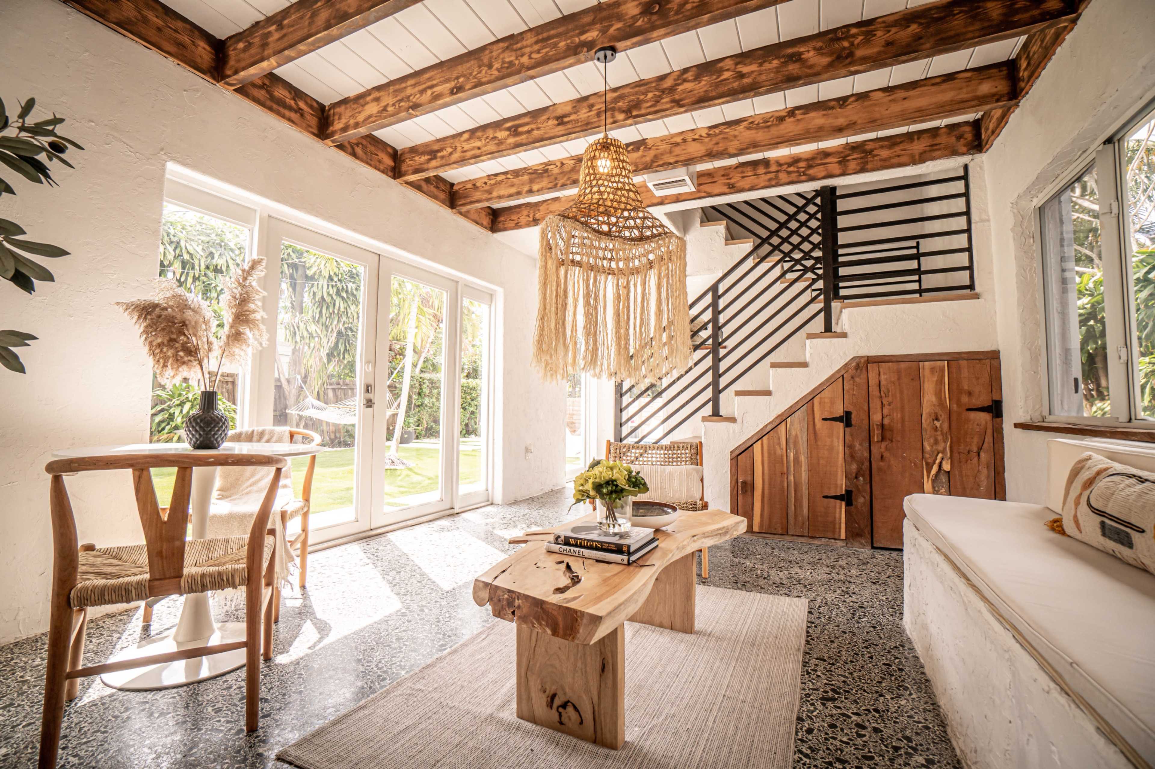 The image shows a bright living room with a wooden table, a woven light fixture, and a staircase leading to an upper level.