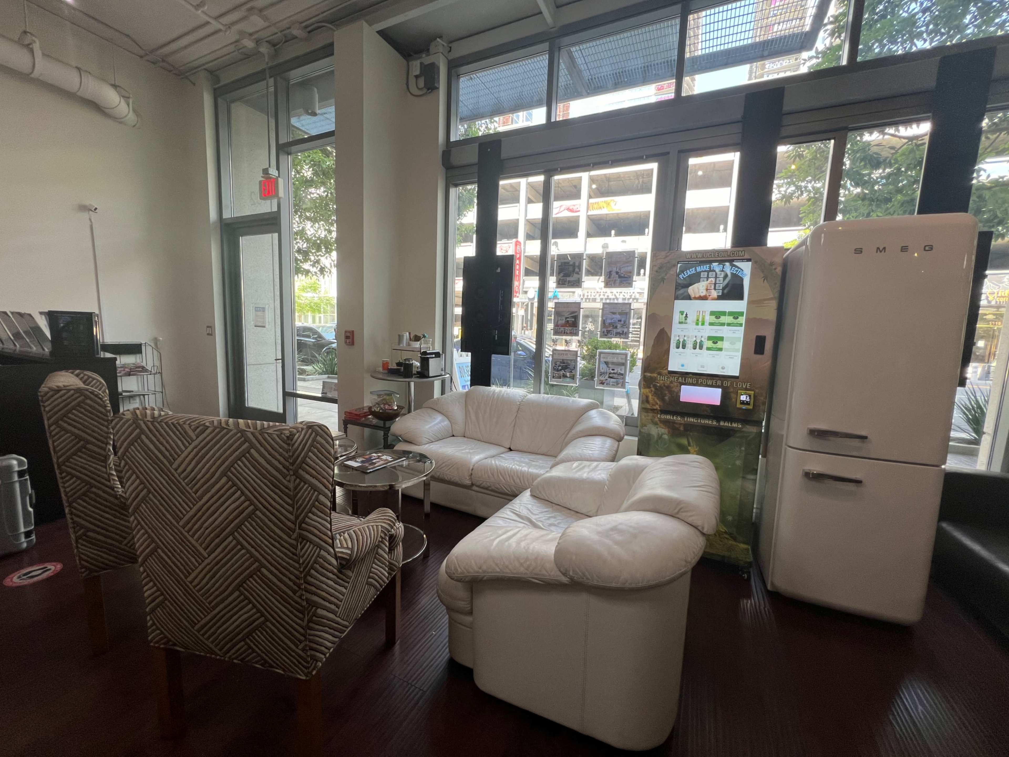 The image shows a modern interior space with a white leather sofa and armchairs, a retro-style refrigerator, and large windows overlooking a parking area.
