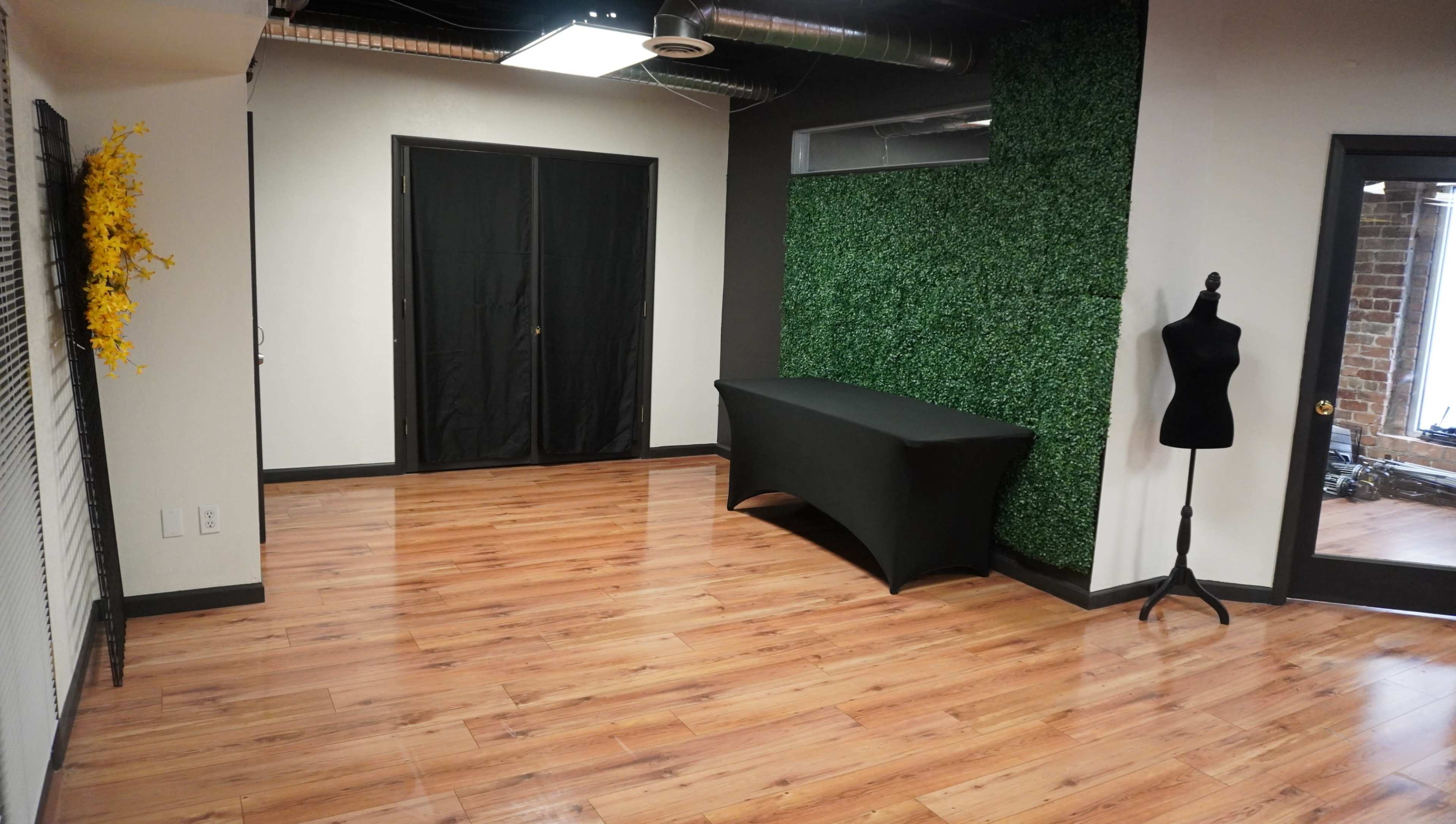 A room with hardwood floors, a black table covered with a cloth, a mannequin, and a green plant wall.