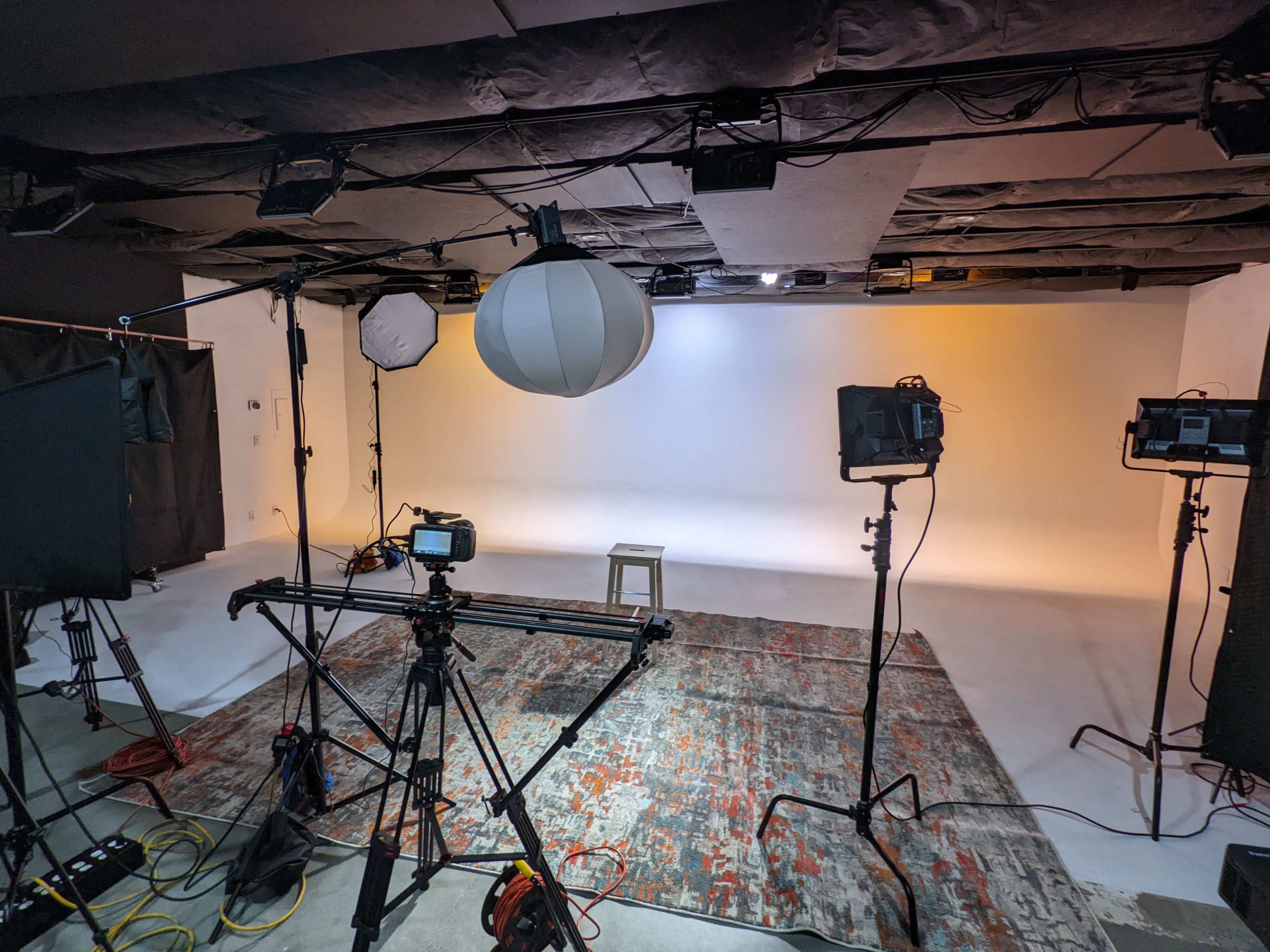 A film studio setup featuring lighting equipment, a camera on a track, and a stool positioned on a patterned rug against a neutral backdrop.