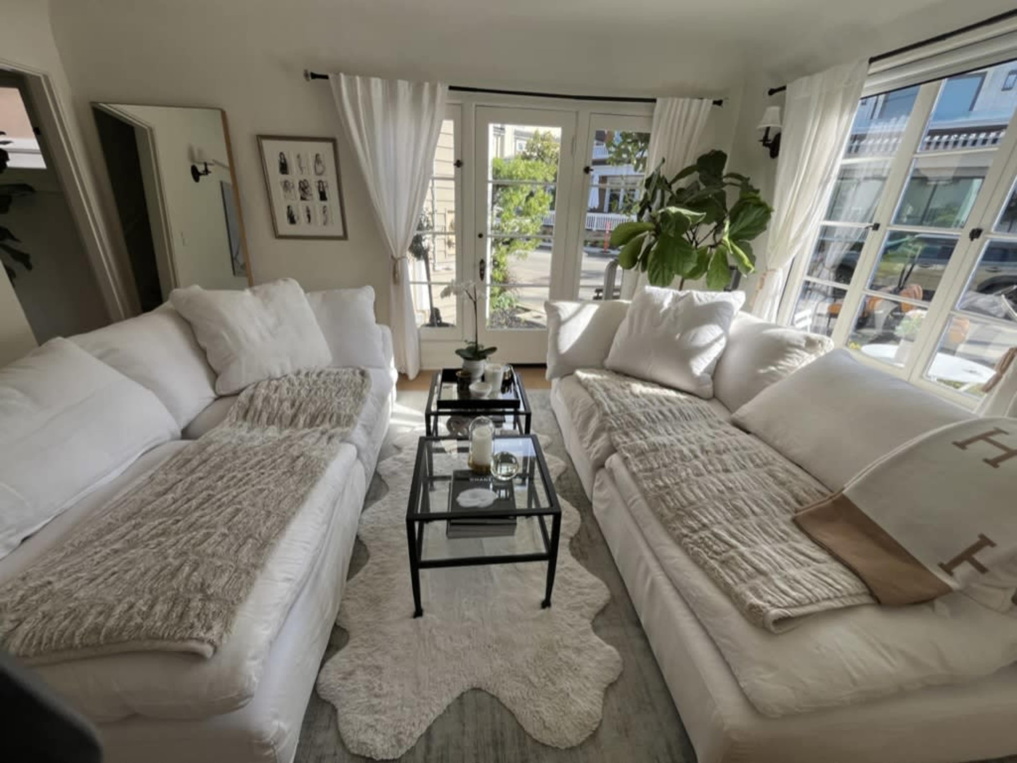 A bright living room features two white sofas facing each other, with a glass coffee table in between and large windows providing natural light.