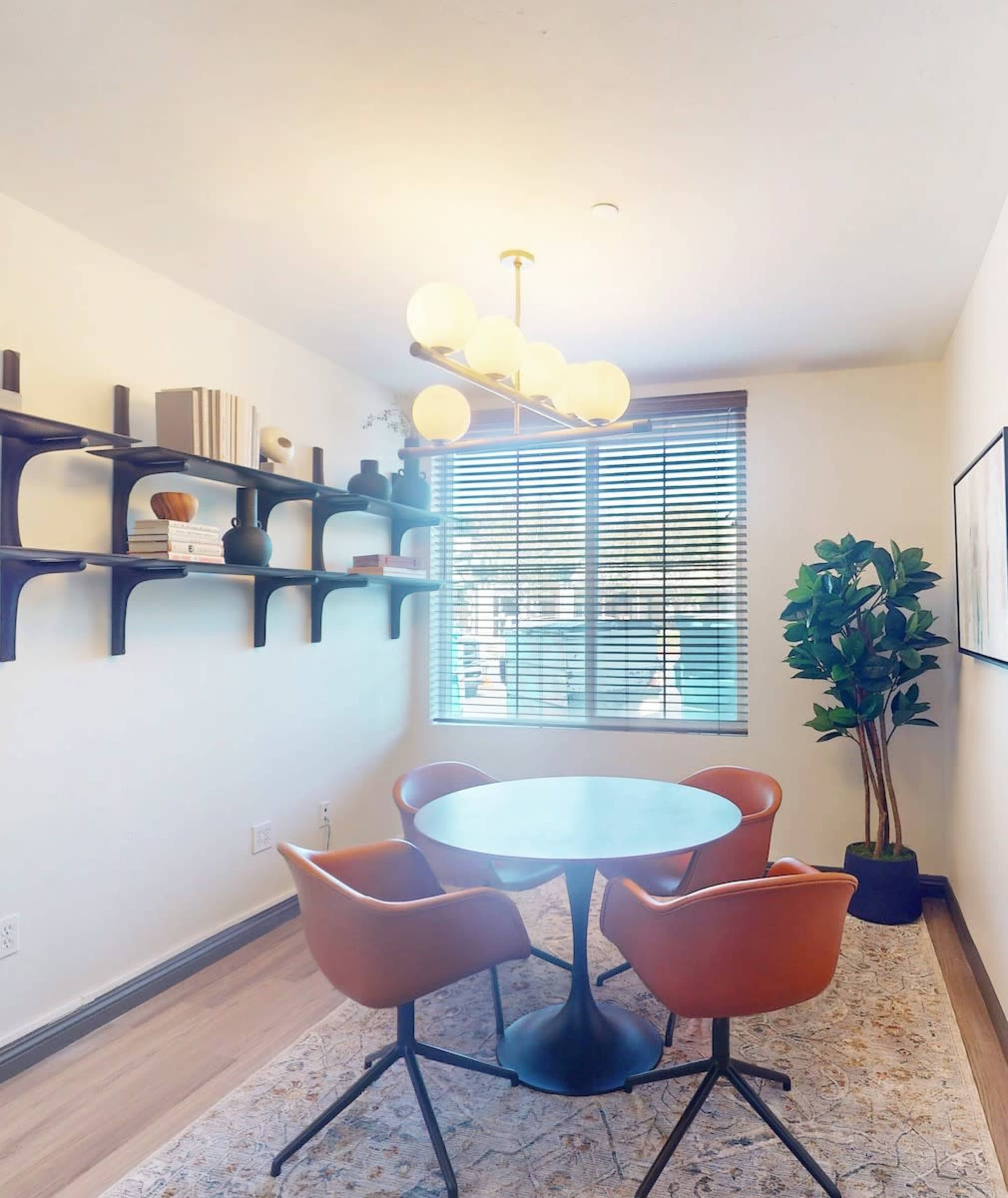 The image shows a modern interior space featuring a circular dining table with two brown chairs, a shelving unit on the wall, and a large window with blinds.