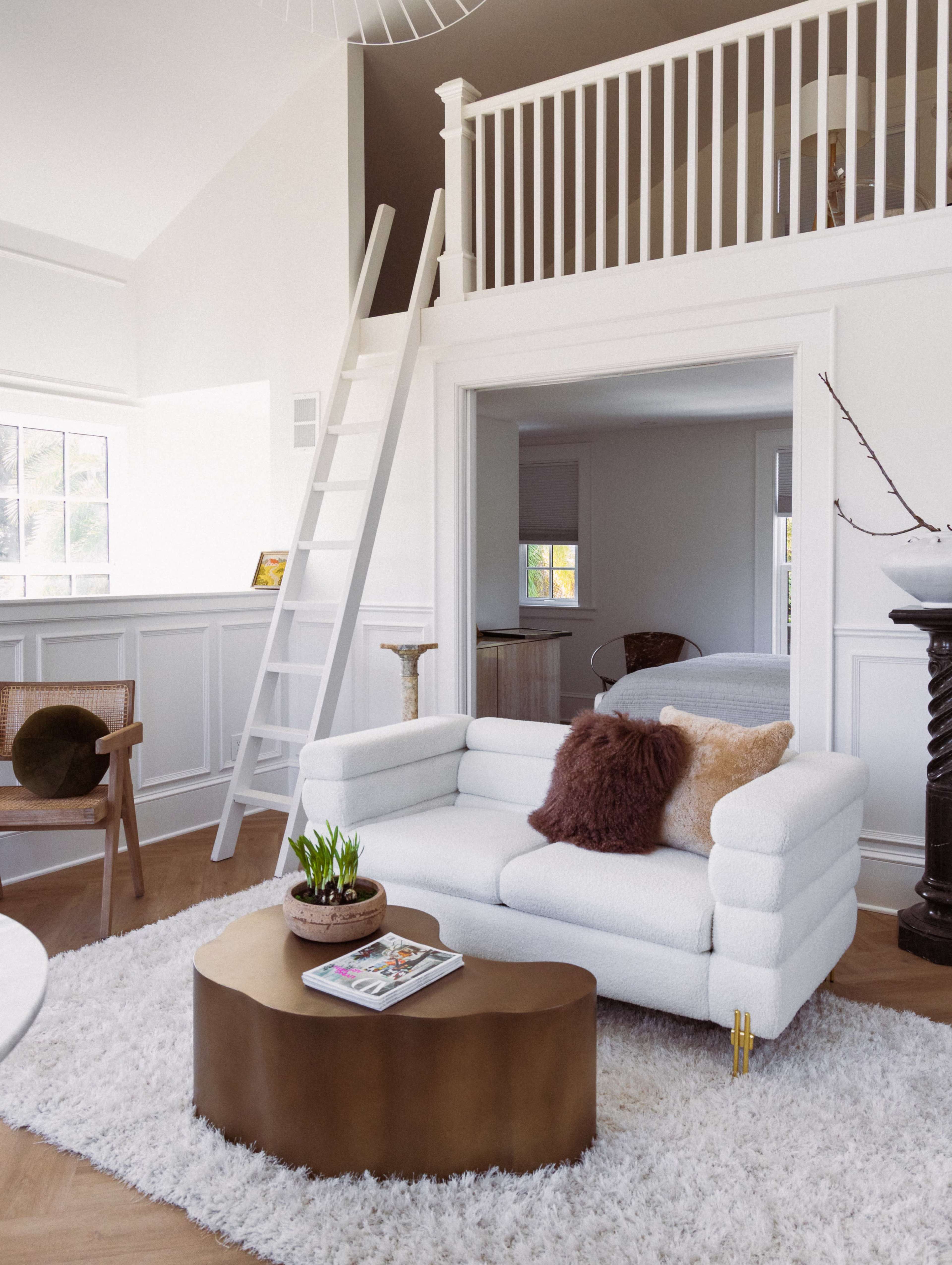The image shows a contemporary living room featuring a white sofa, a round coffee table, and a ladder leading to a loft area.